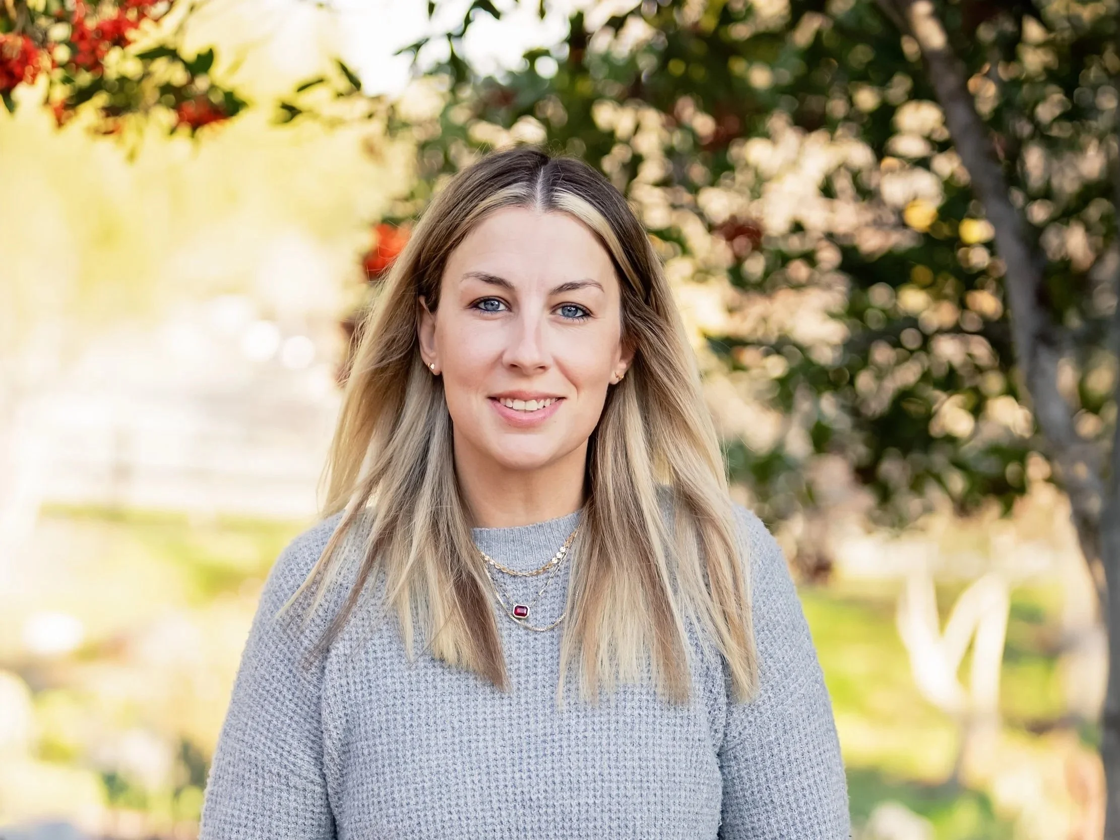 A woman with blonde hair, blue eyes, and light makeup smiling outdoors in front of trees and sunlight.