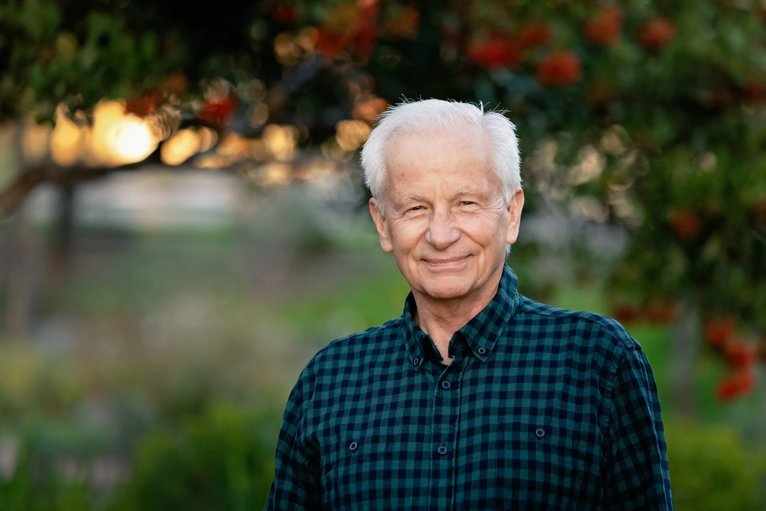 An elderly man with white hair and a slight smile stands outdoors in front of a blurred background of trees and plants during daytime.
