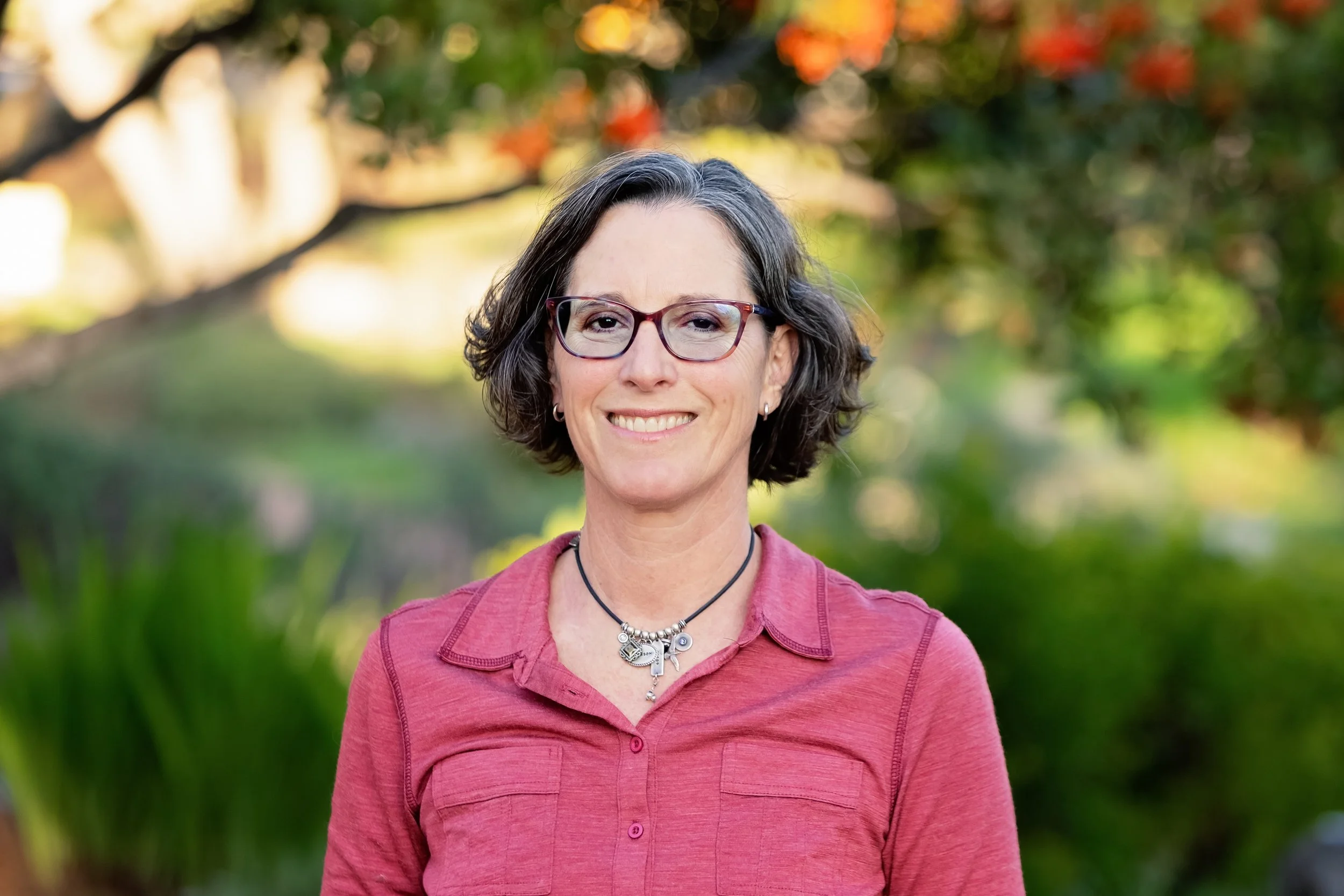 A woman with short dark hair, glasses, and wearing a pink shirt outdoors in front of greenery and trees with fall-colored leaves.