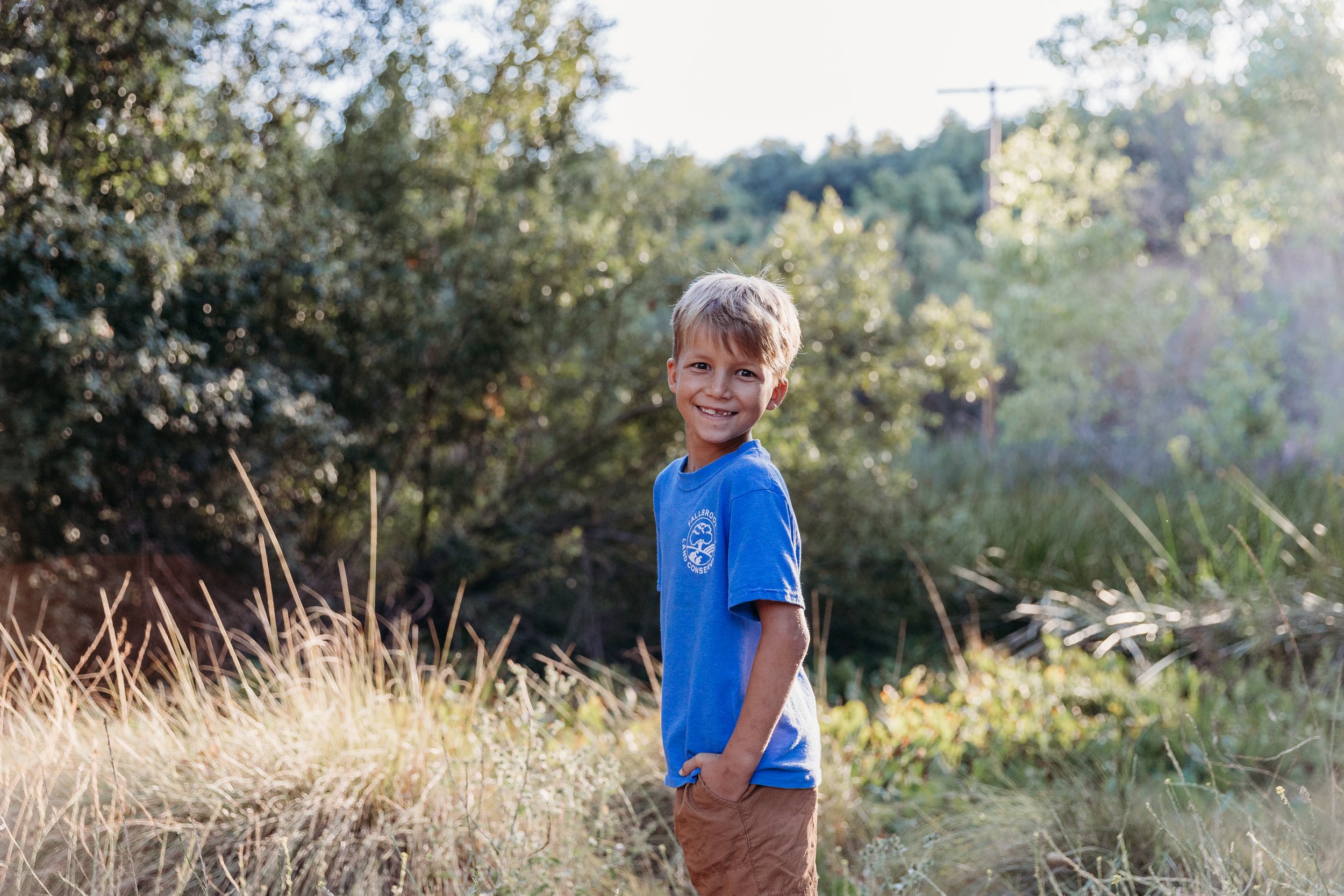 A young boy smiling outdoors, standing in a grassy area with trees in the background, wearing a blue t-shirt and brown shorts.