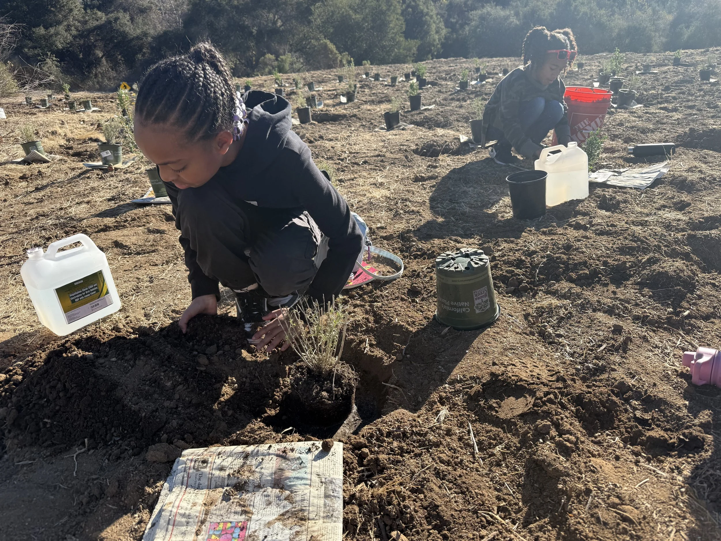 Two women planting small plants in a field with rows of small potted plants, dirt, and gardening supplies around them.