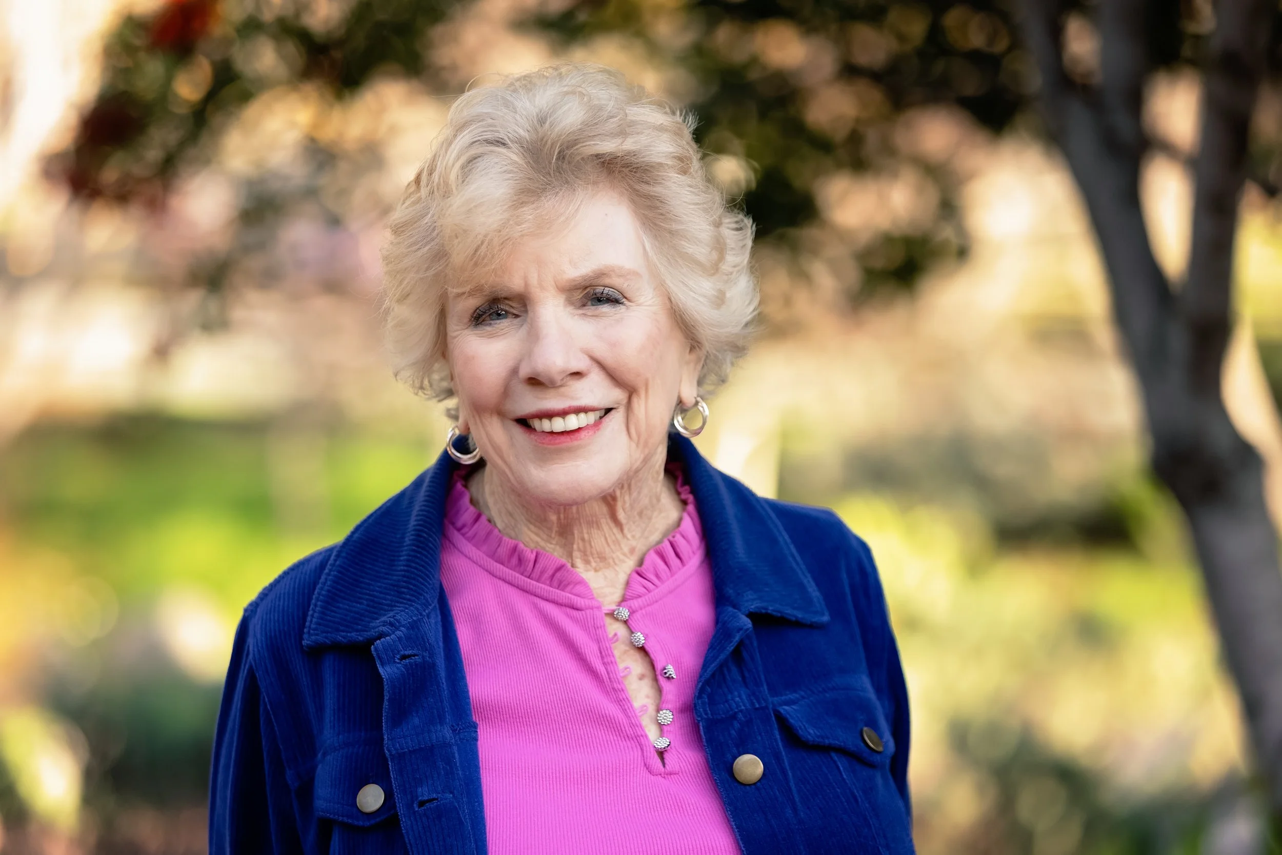 Older woman with curly gray hair smiling outdoors, wearing a pink shirt and blue jacket, with blurred trees in the background.