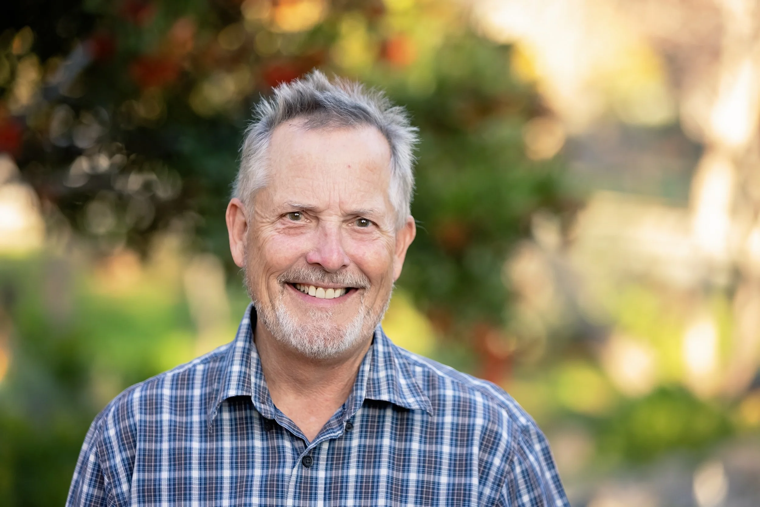 A smiling older man with gray hair and a beard, wearing a blue checked shirt, outdoors with blurred green and autumn-colored trees in the background.