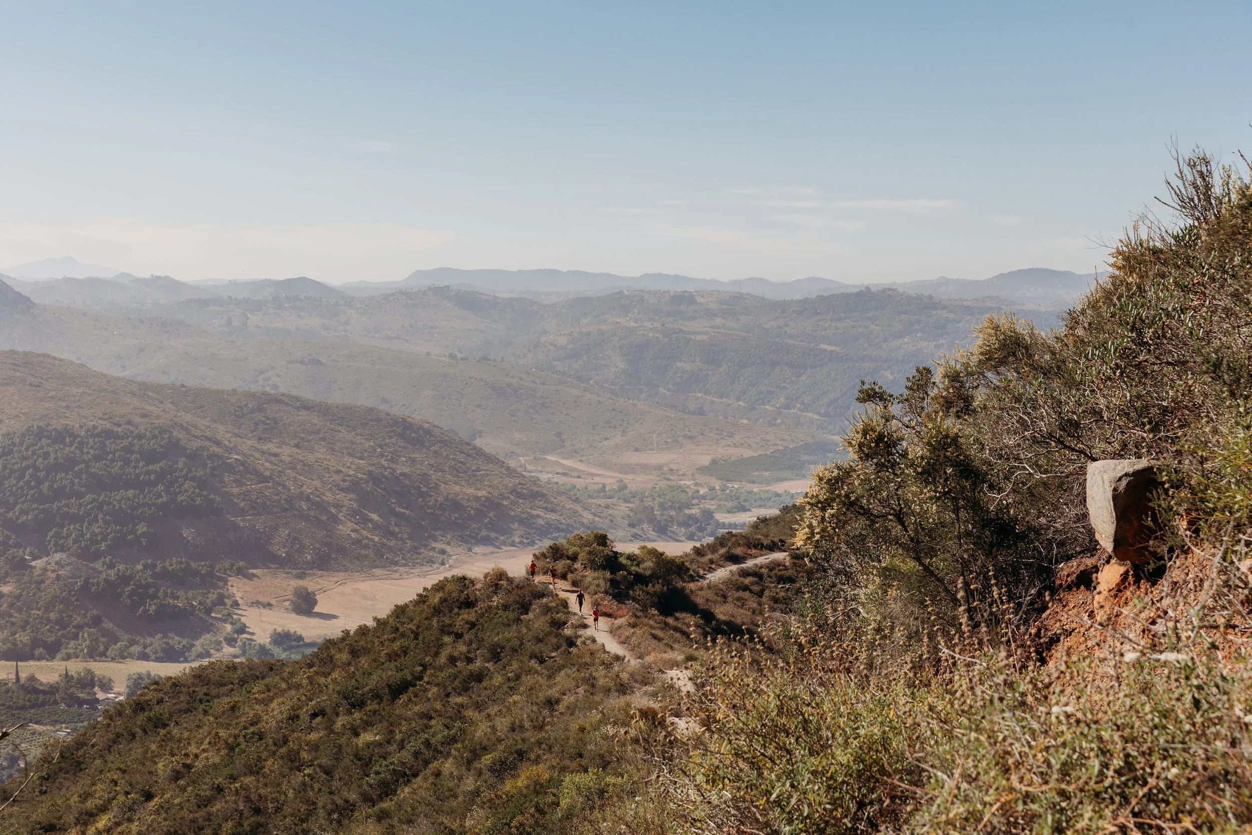 Hikers walking along  the Monserate Mountain trail surrounded by dry bushes and sparse trees, with a distant view of rolling hills and valleys under a clear blue sky.