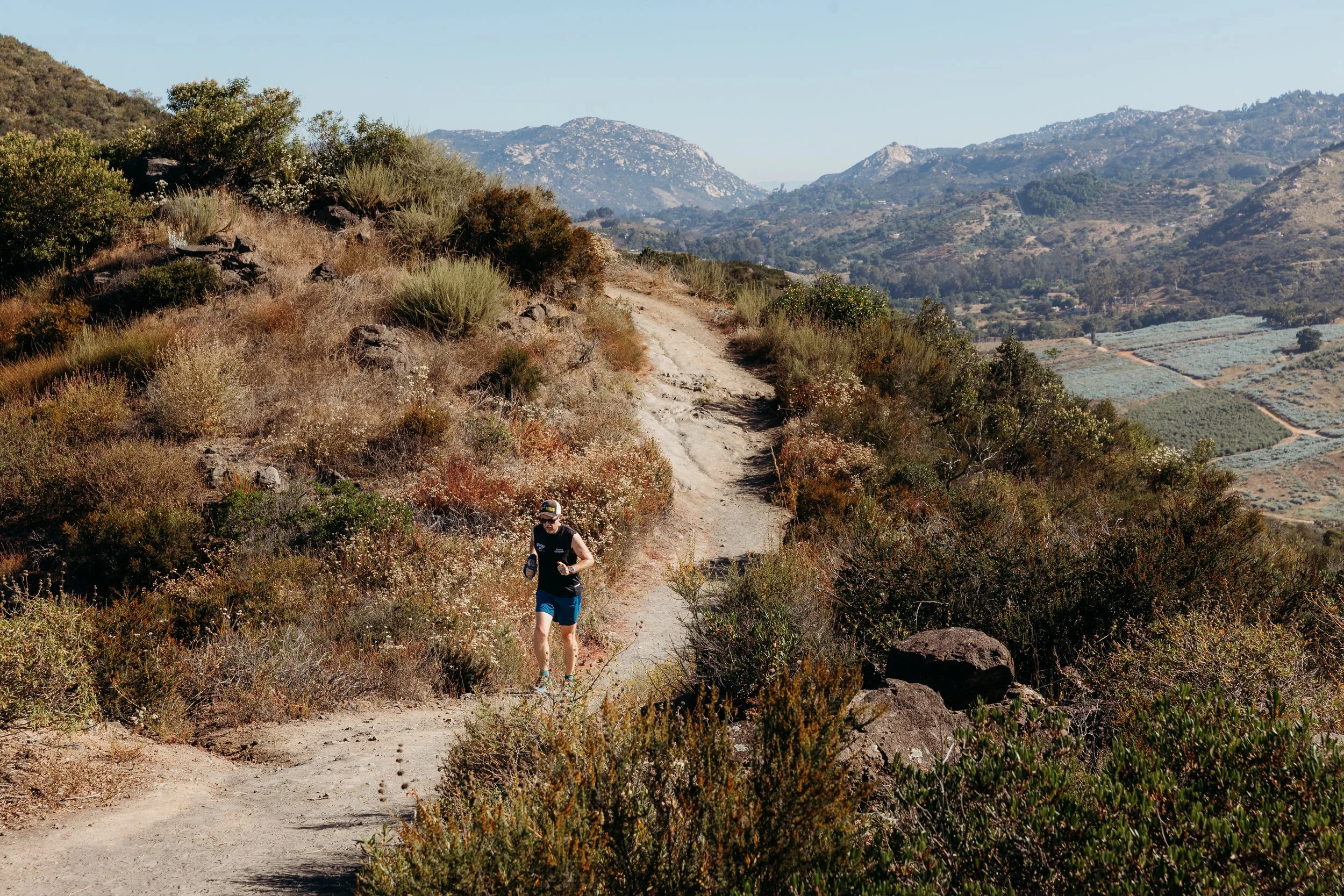 A man running on a dirt trail in a hilly, semi-arid landscape with shrubs and mountains in the background.