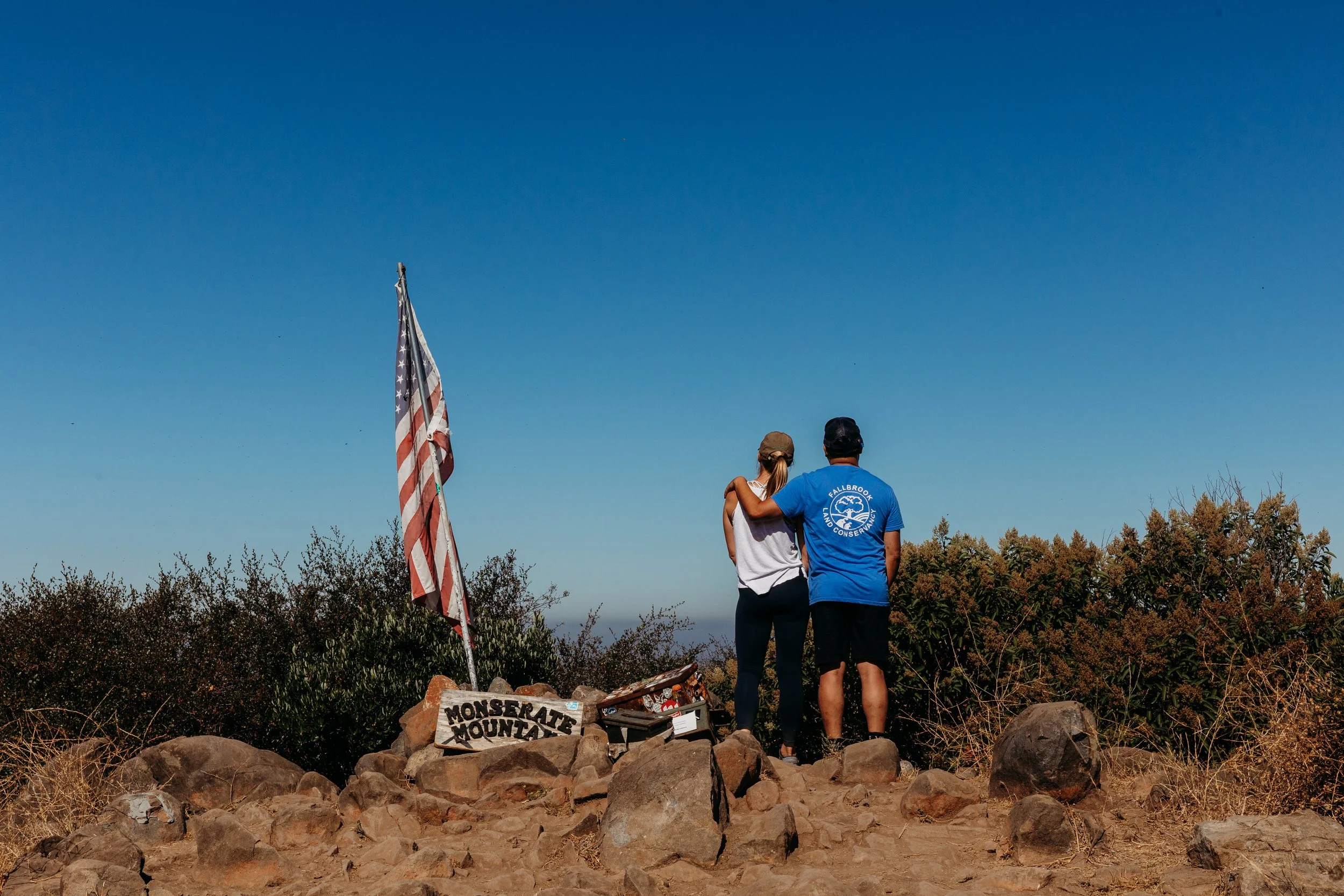 A man and woman stand together on a mountain summit, with their arms around each other, looking at the view. They are near a U.S. flag on a pole atop Monserate Mountain.