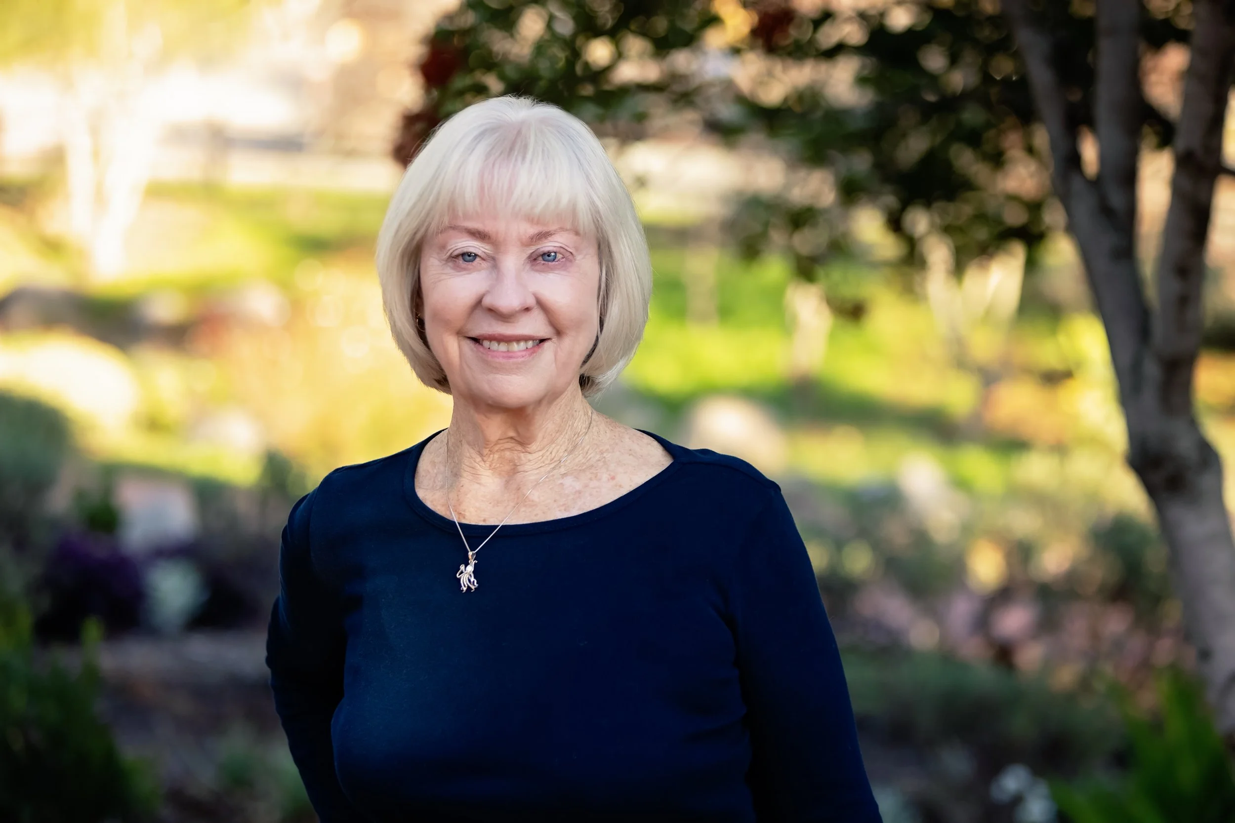 An elderly woman with short blonde hair smiling outdoors, wearing a navy blue top and a silver necklace, with trees and greenery in the background.