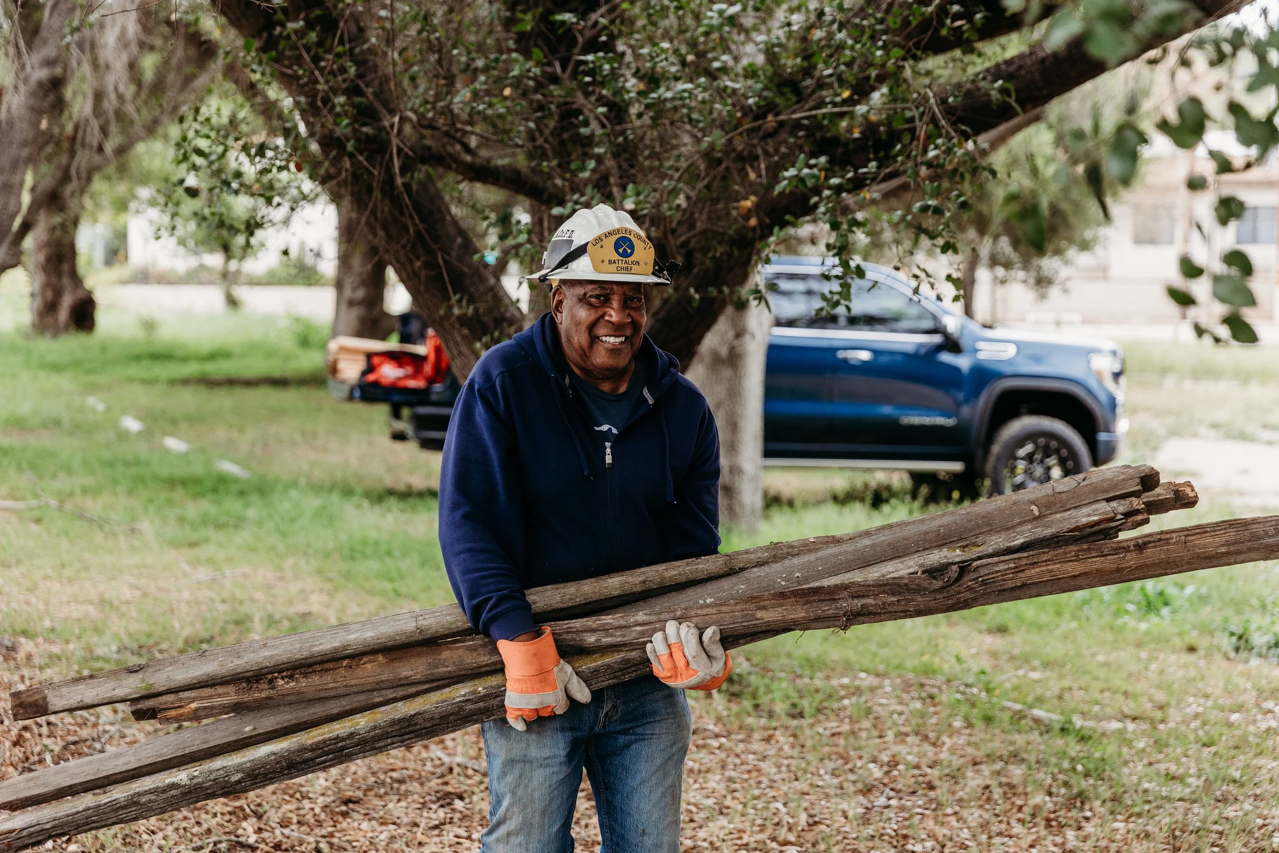 A man wearing a helmet and gloves is holding a long wooden log, standing outdoors near a tree with a blue vehicle in the background.