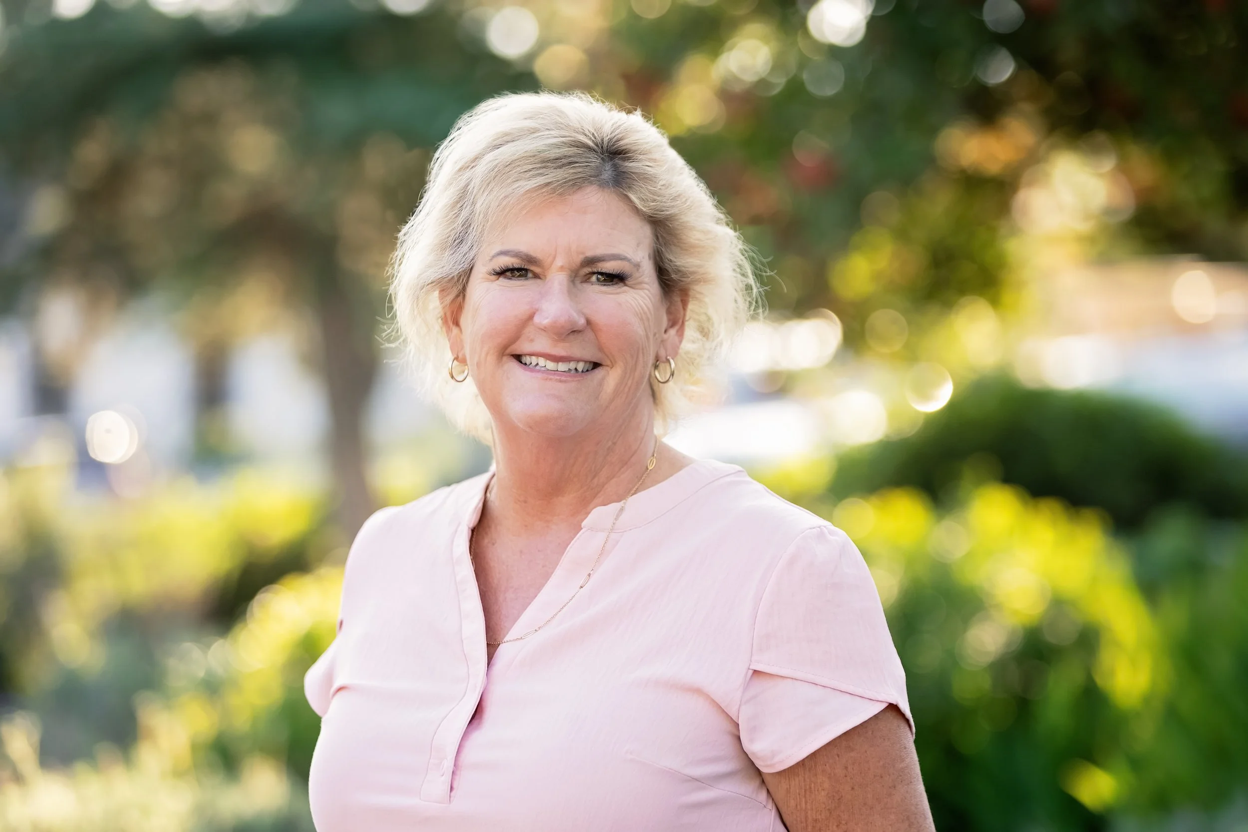 Smiling elderly woman with short blonde hair standing outdoors with trees and greenery in the background.