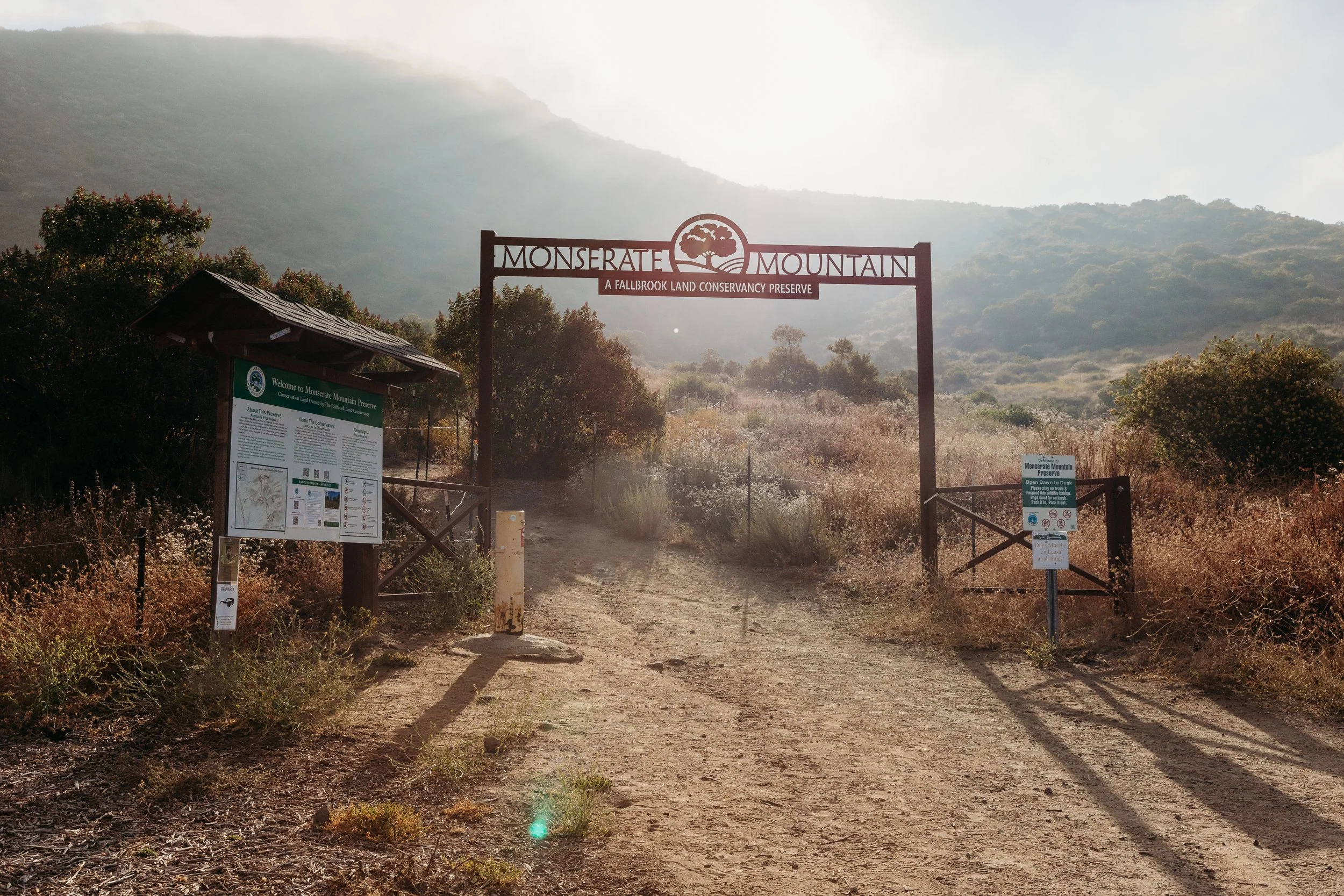 Entrance gate to Monserrate Mountain Preserve with informational signs and a dirt road leading into the preserve, surrounded by dry grasses and bushes, with mountains in the background and sunlight shining through haze.