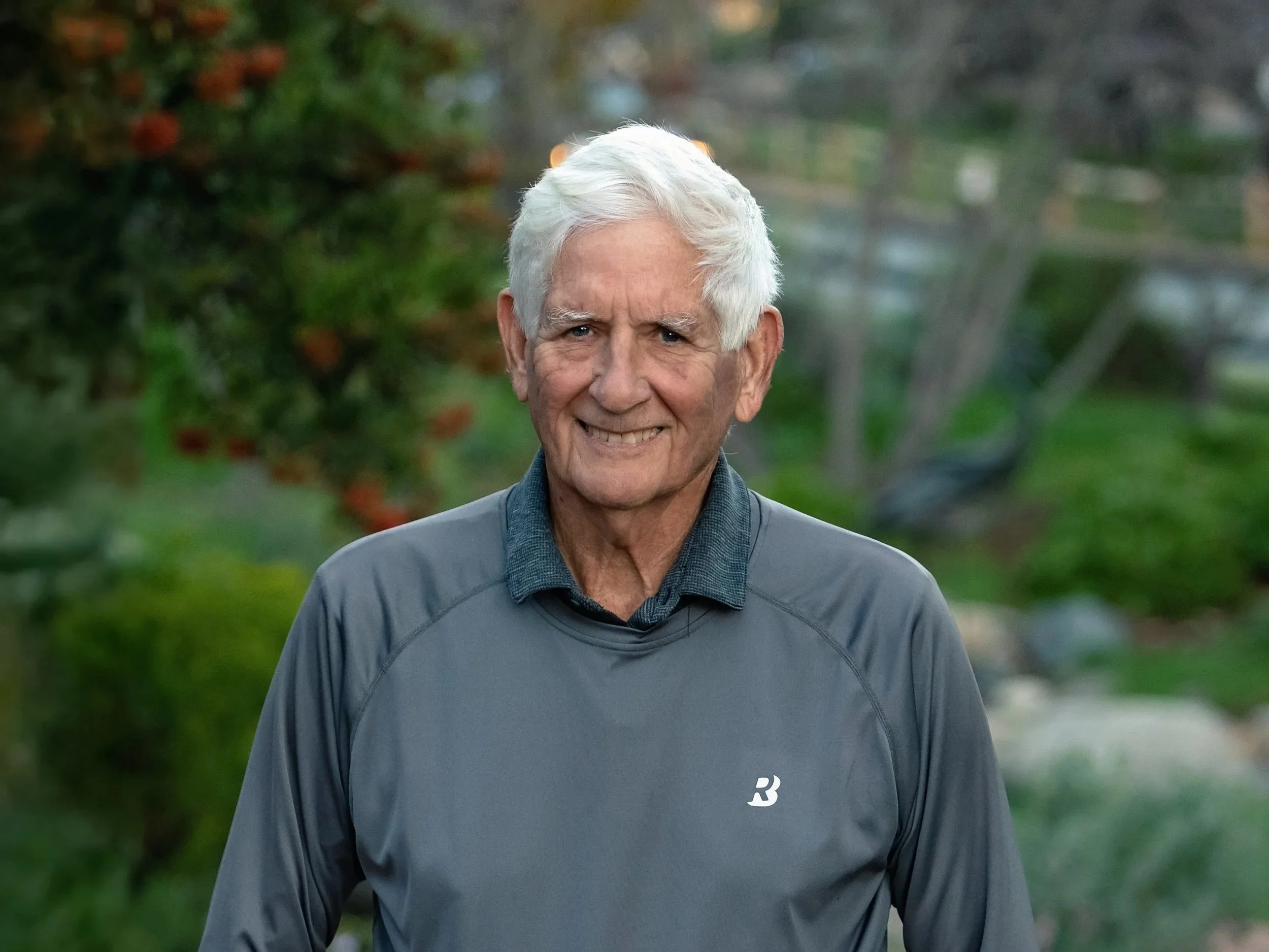 An elderly man with white hair smiling outdoors in a park, wearing a gray athletic jacket.