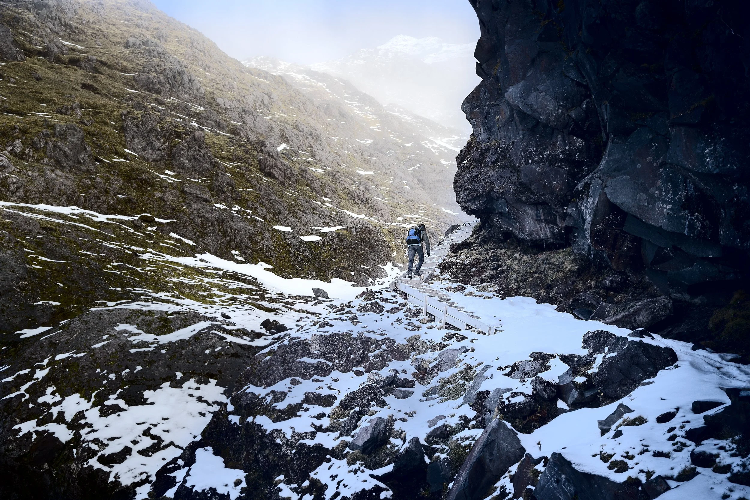 A person with a backpack hiking on a snowy trail through a rocky mountain pass with steep cliffs and snow patches.