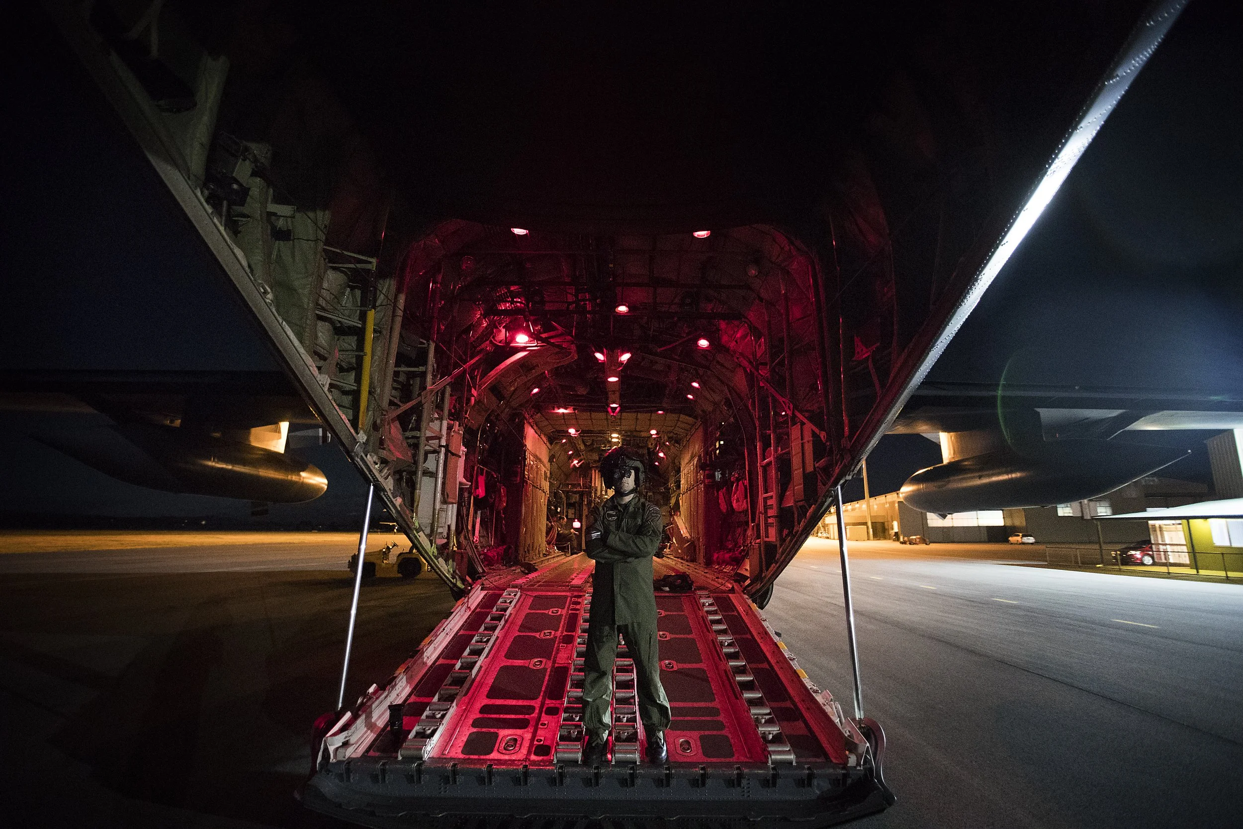 A military personnel in uniform standing inside a cargo plane with its rear ramp open, illuminated by red lights, on an airport tarmac at night.