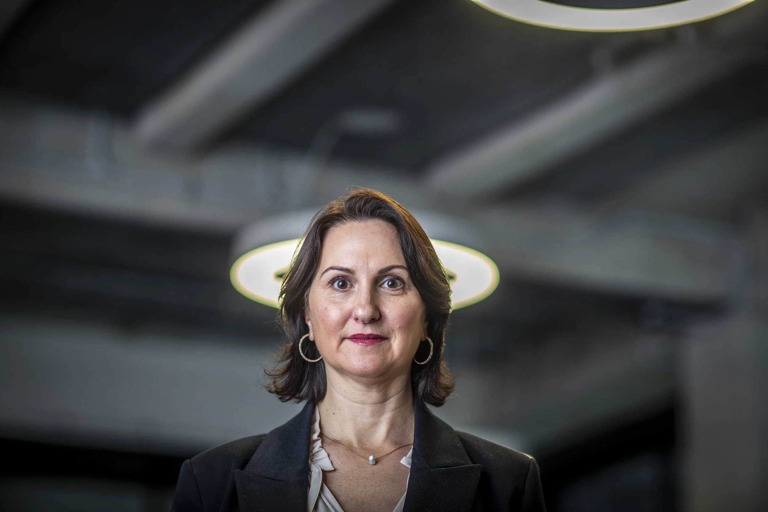 A woman with shoulder-length dark hair, wearing a black blazer and hoop earrings, standing indoors with modern ceiling lights in the background.