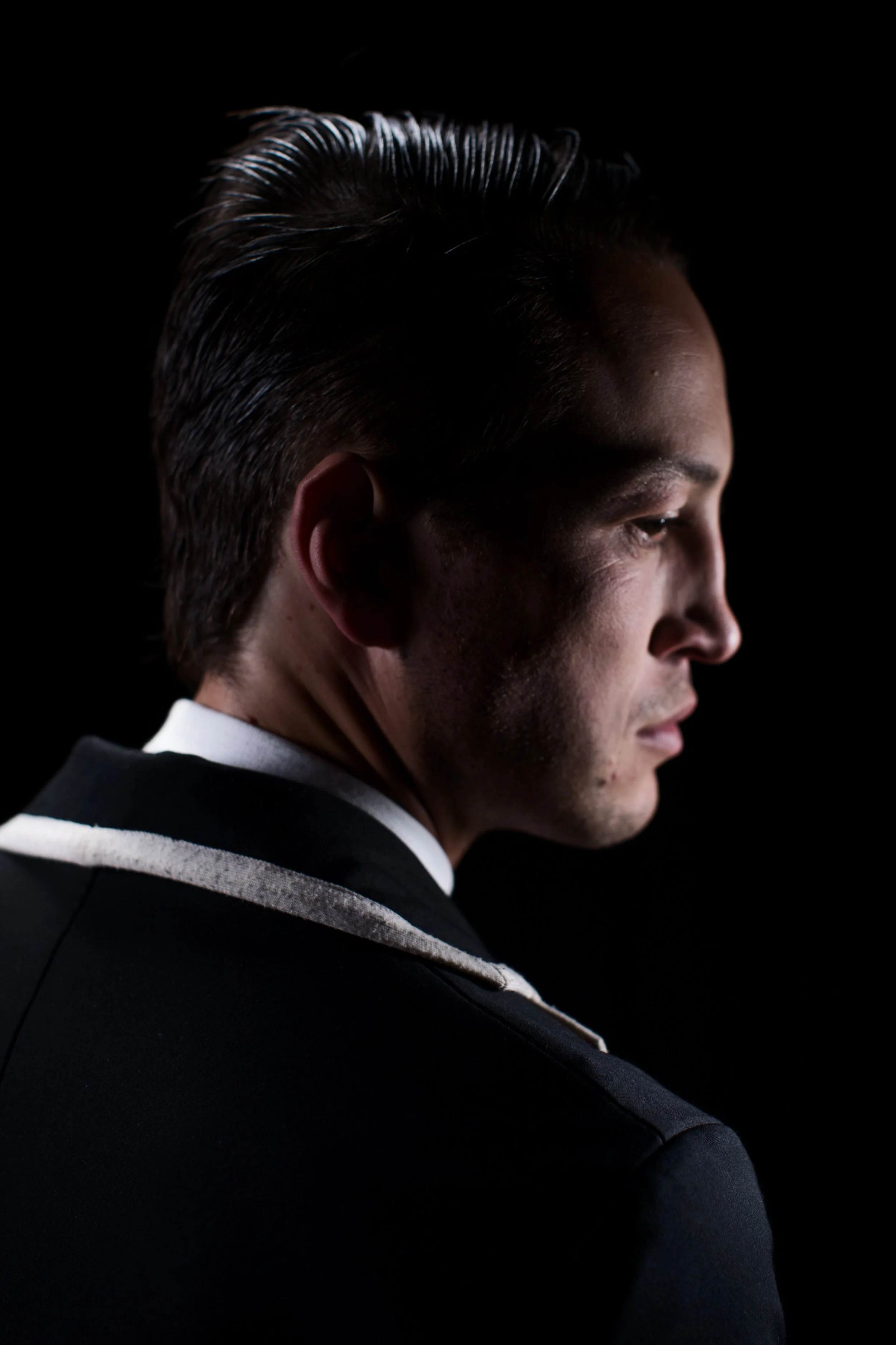 Side profile of a man with slicked-back hair, wearing a dark suit with a white collar, against a black background.