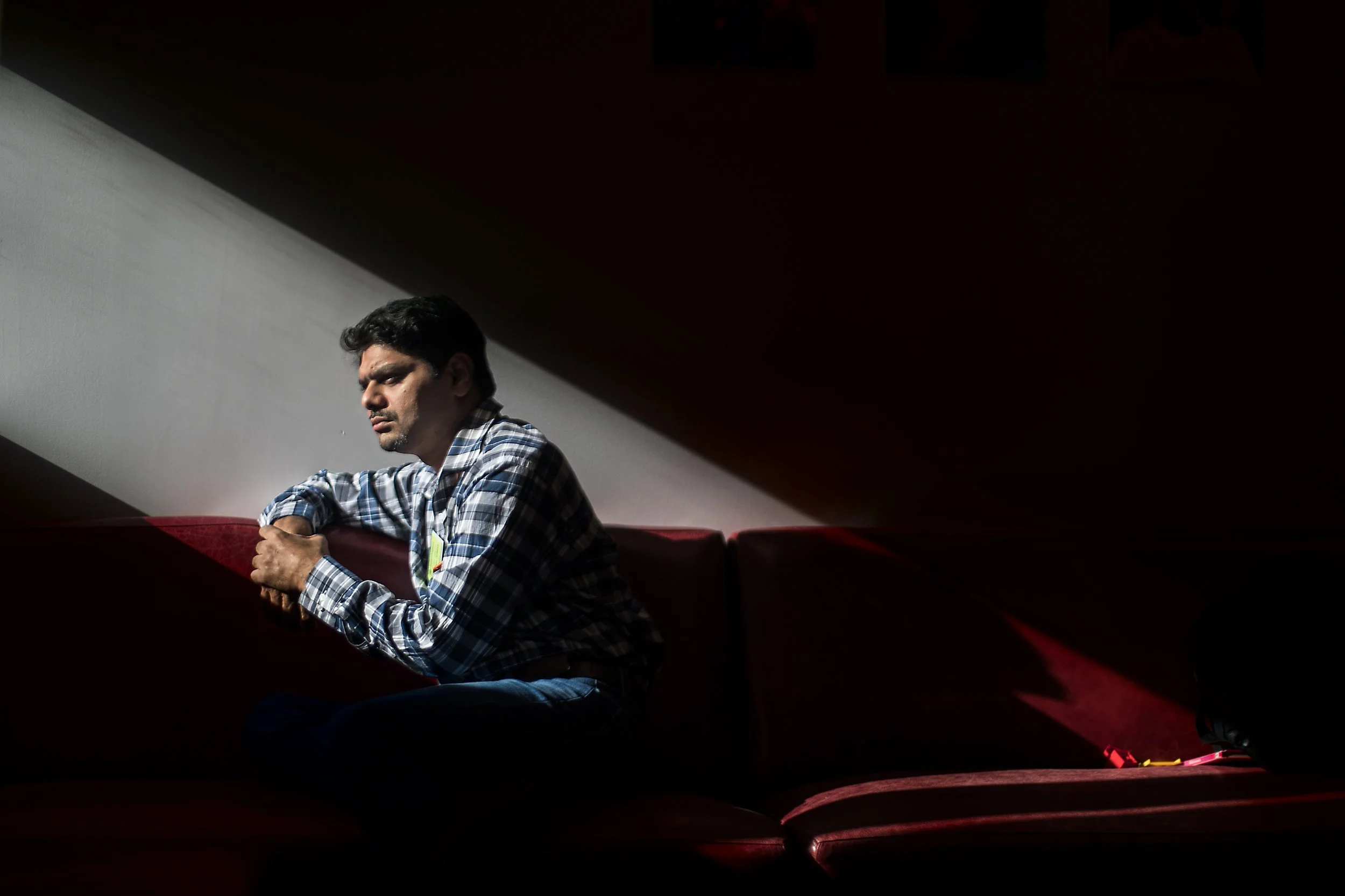 A man sitting alone on a red couch, with one arm resting on the back of the couch, appears pensive in a dimly lit room with a sharp light illuminating part of his face and shirt.