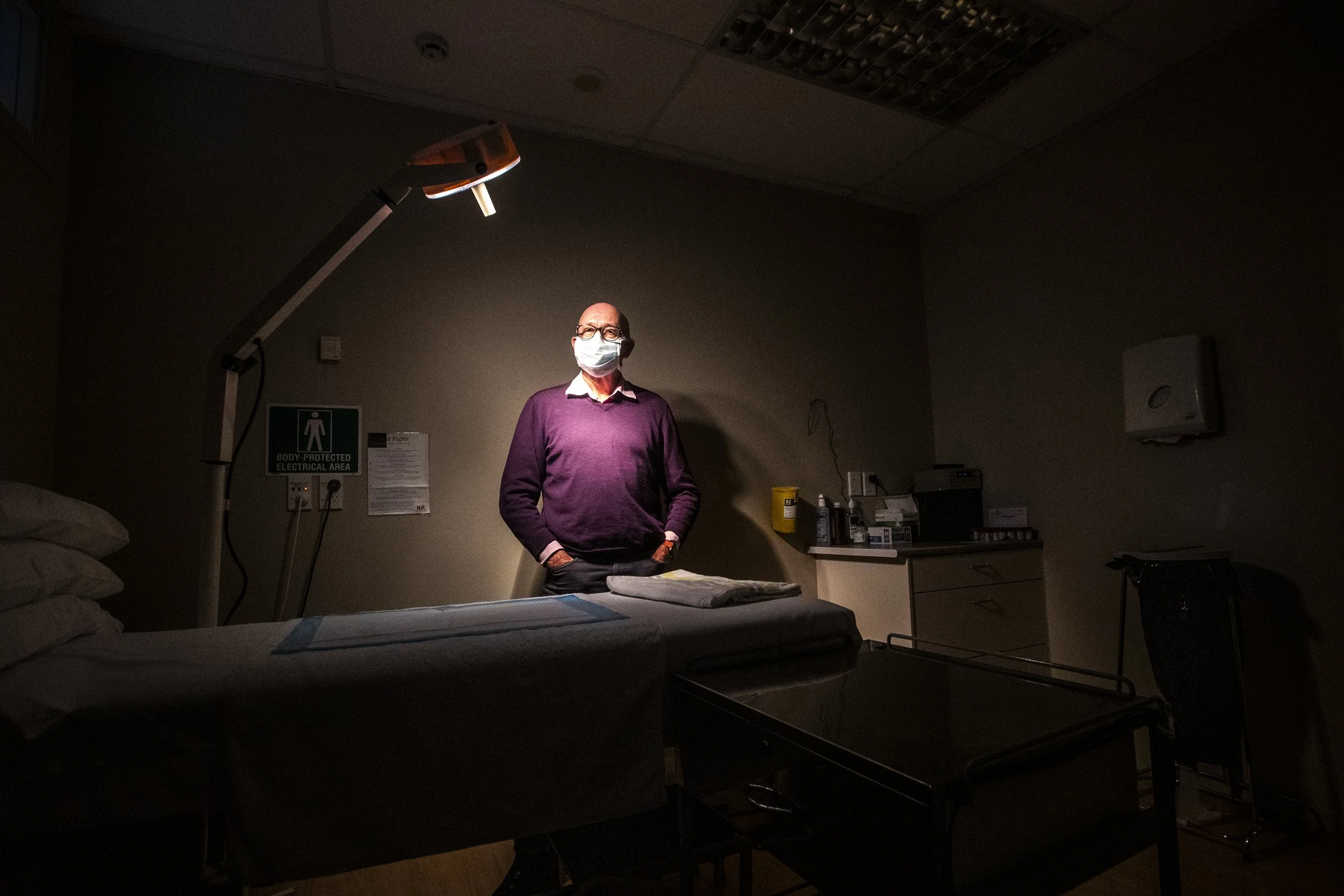 A man wearing glasses and a face mask standing in a dimly lit hospital room, with medical equipment and a hospital bed visible.