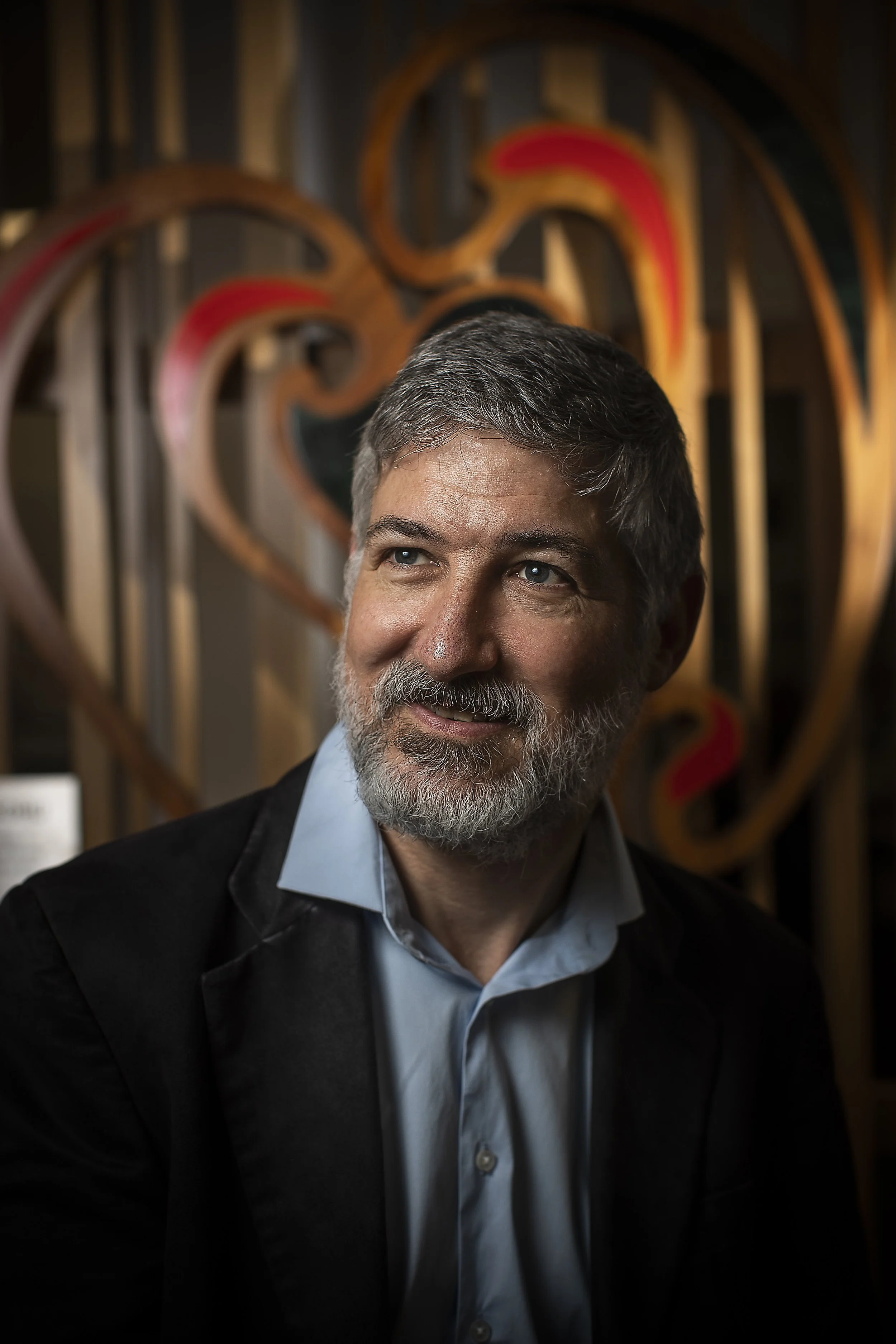 A man with gray hair and beard, wearing a gray shirt and black blazer, looking to the side with a slight smile, standing in front of a wooden heart-shaped art piece with red accents.