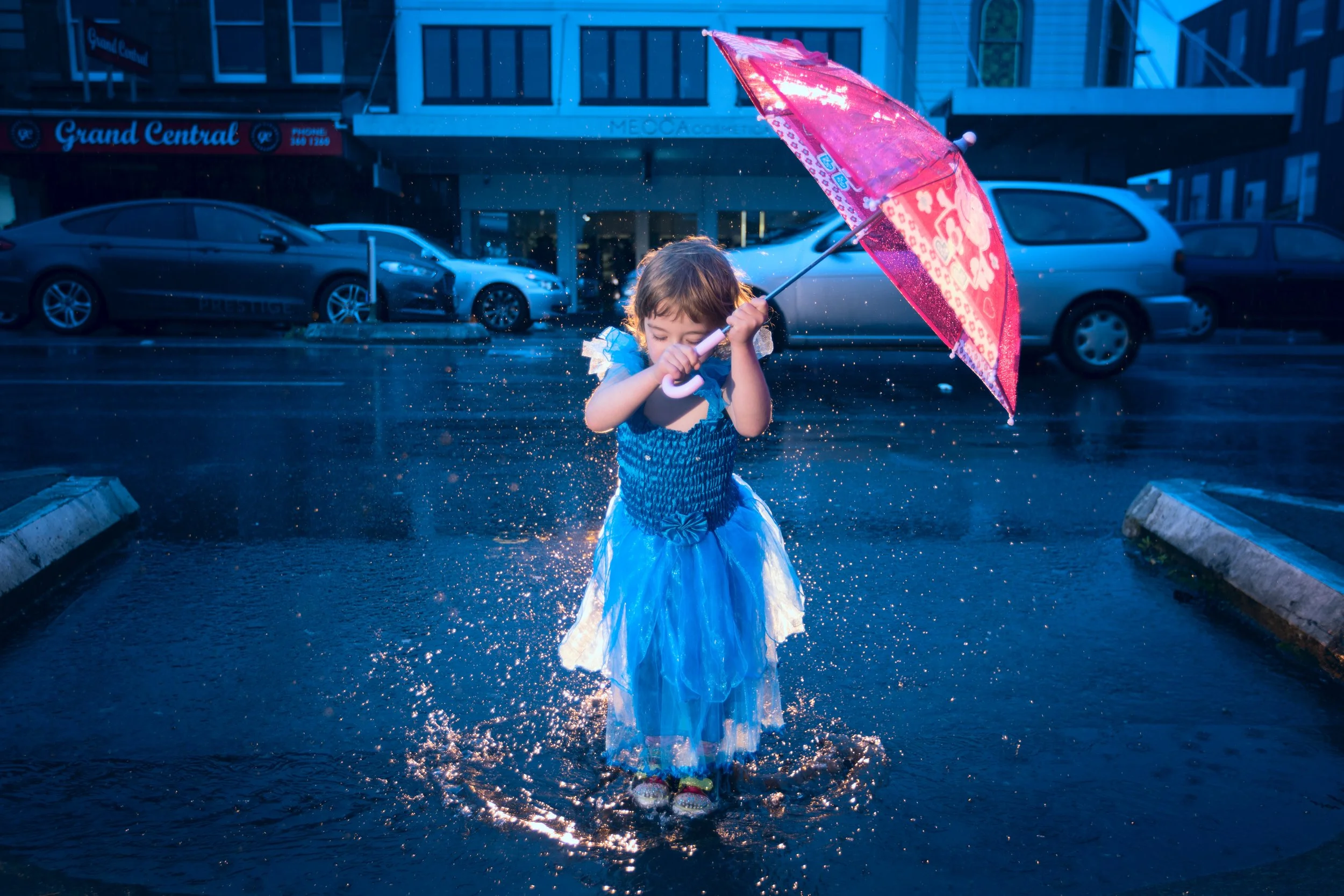 A young girl in a blue dress playing in a puddle on a rainy day, holding a pink umbrella with a floral pattern.
