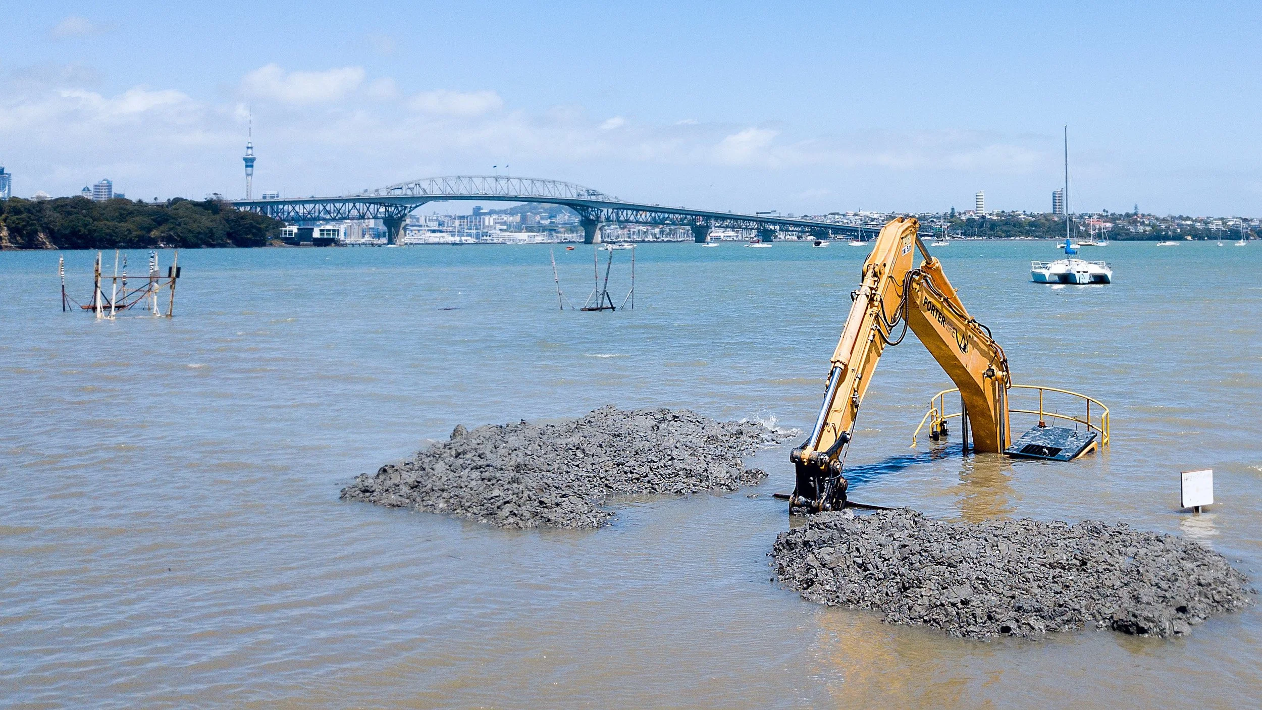 A construction excavator partially submerged in water, working on site near a river with boats and a city skyline in the background.