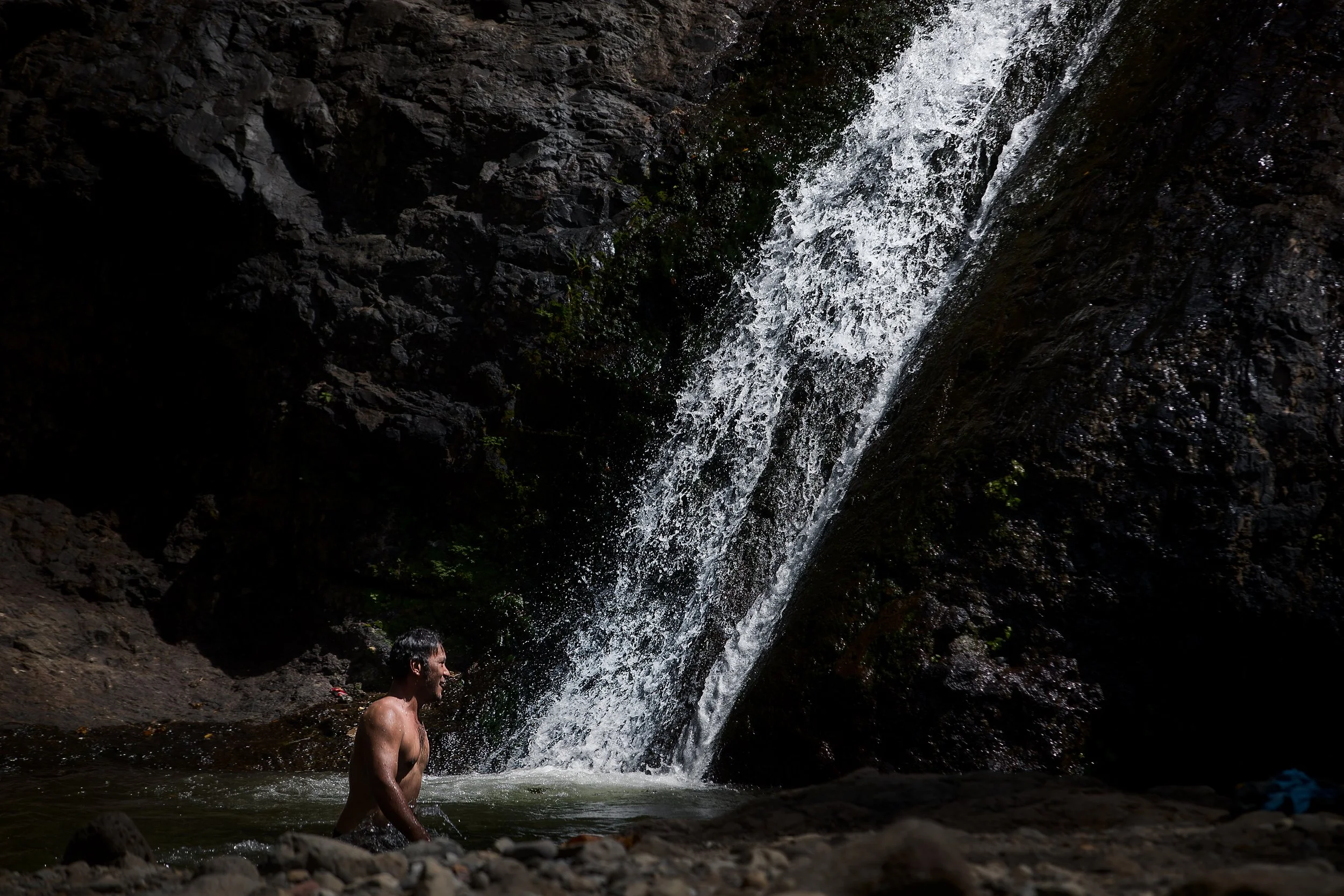A man standing in a small pool of water near a waterfall surrounded by dark rocks.