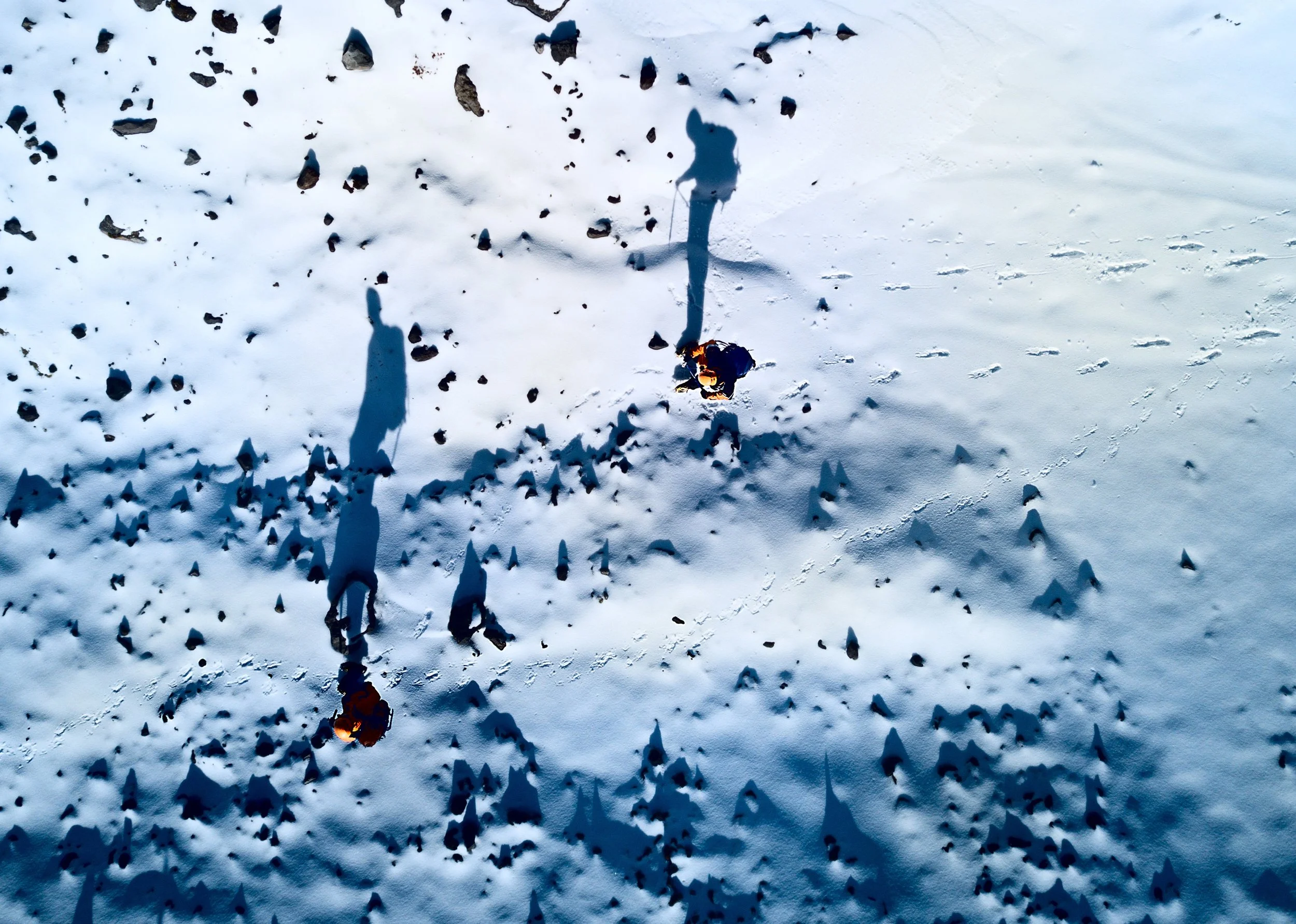 Two people in winter clothing walking in snow, casting long shadows on the snow-covered ground, with scattered rocks and footprints visible.