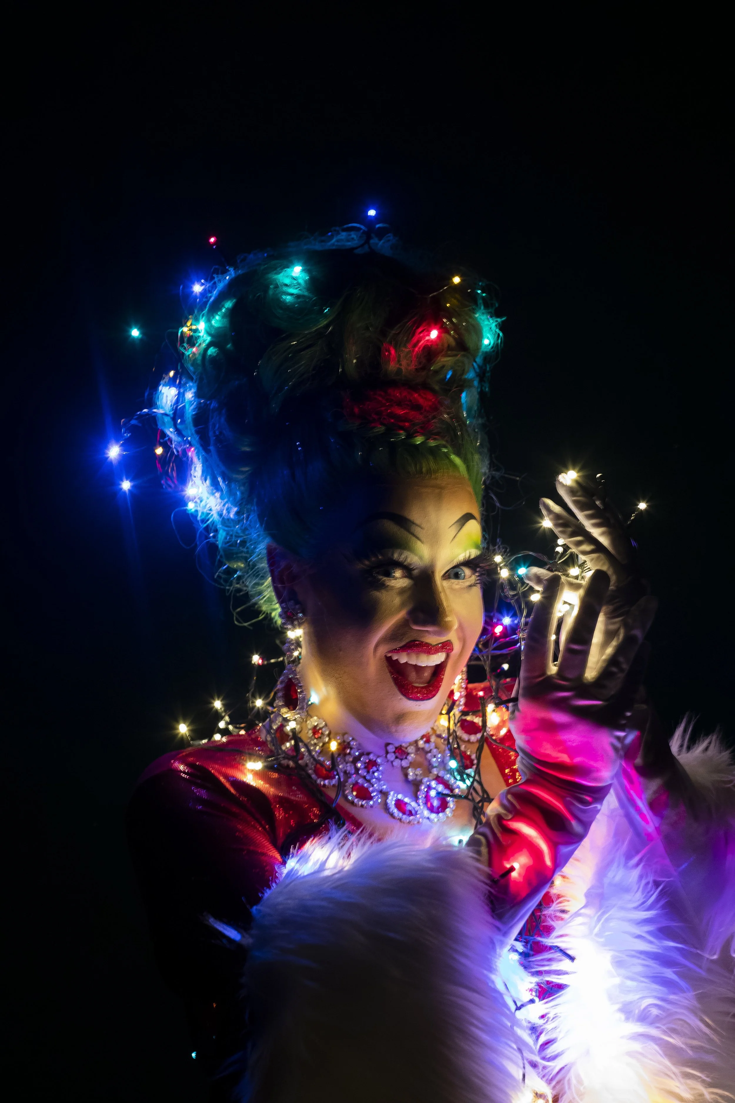 A performer dressed in festive holiday attire, wearing colorful Christmas lights in her hair and jewelry, with a wide smile, set against a dark background.