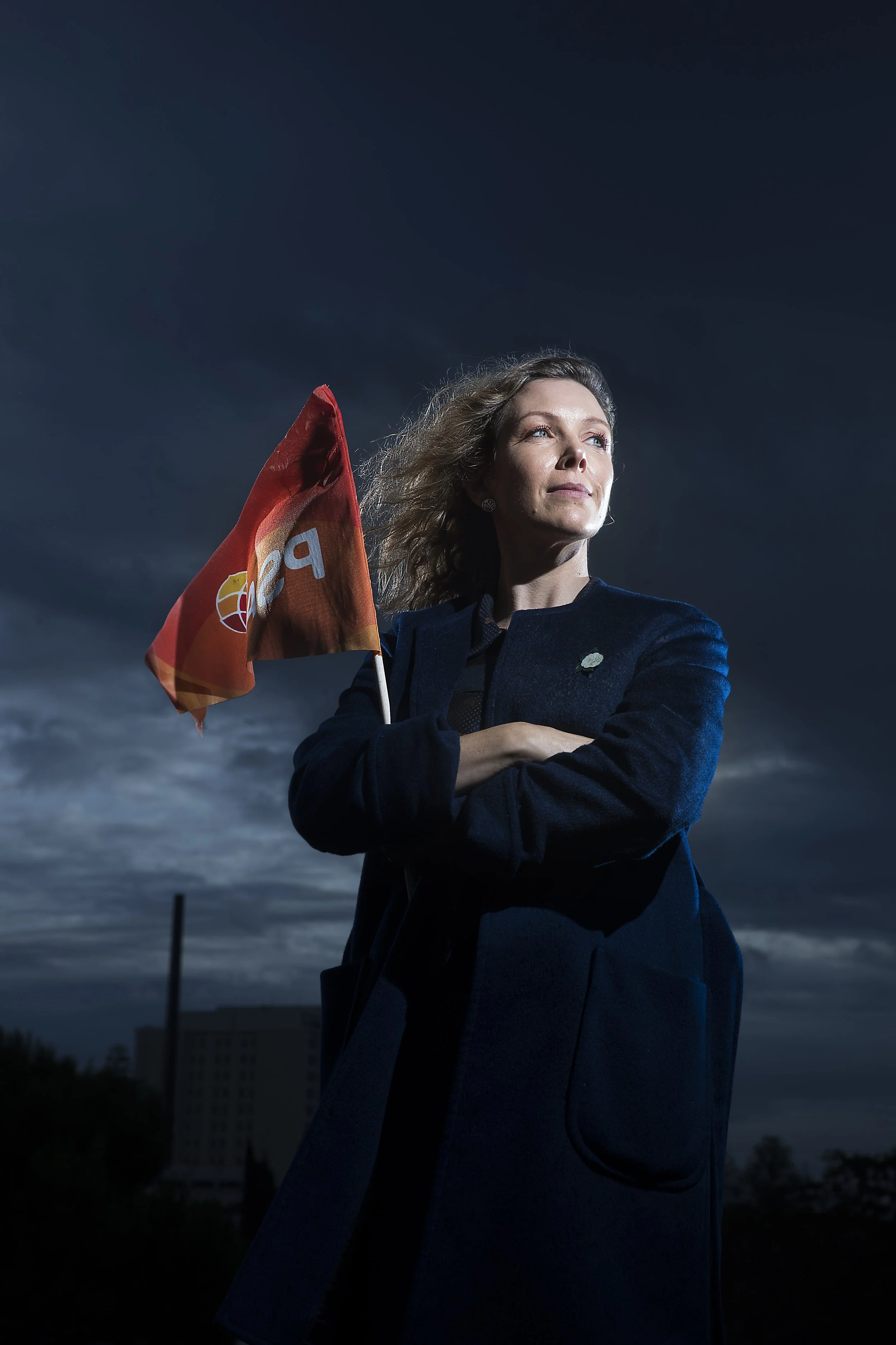 A woman with wavy hair holding a flag, standing outdoors during dusk or night with a dark sky behind her.