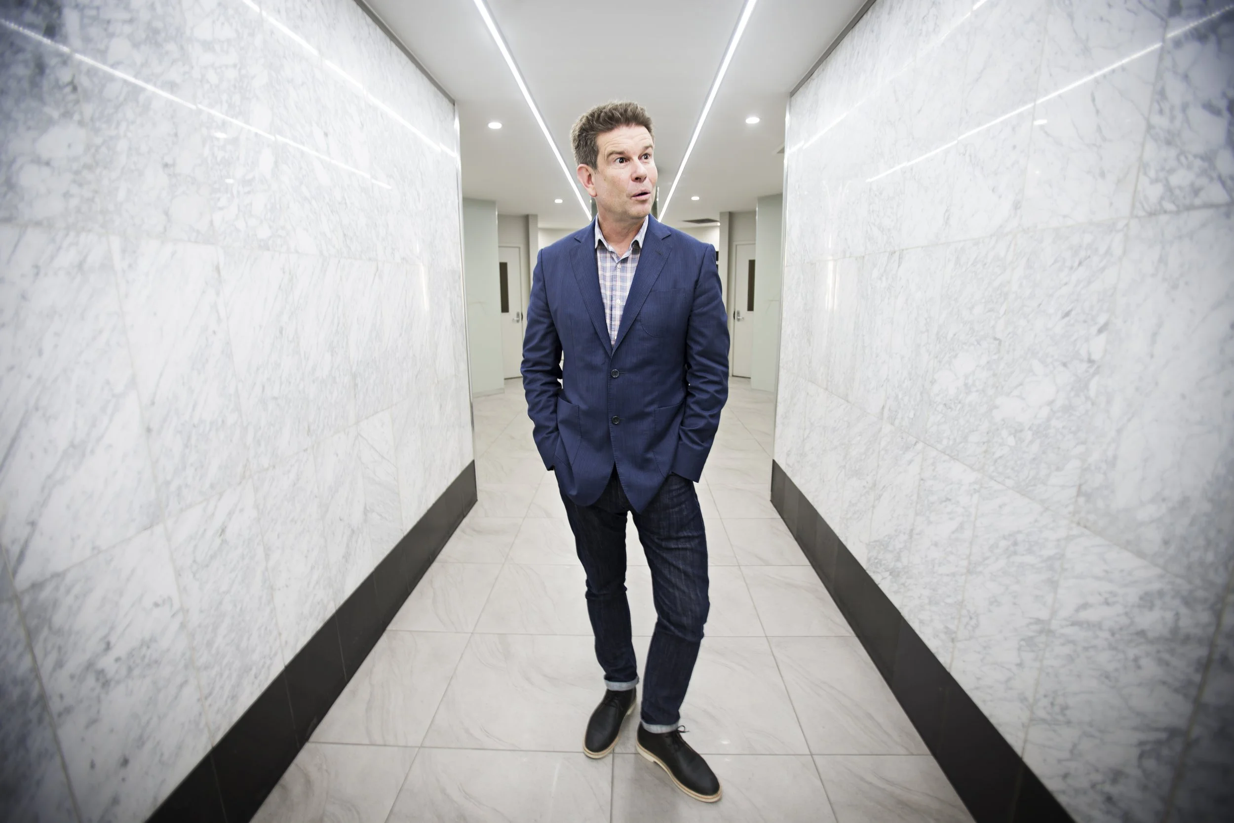 A man in a blue blazer and jeans walking in a brightly lit hallway with marble walls.