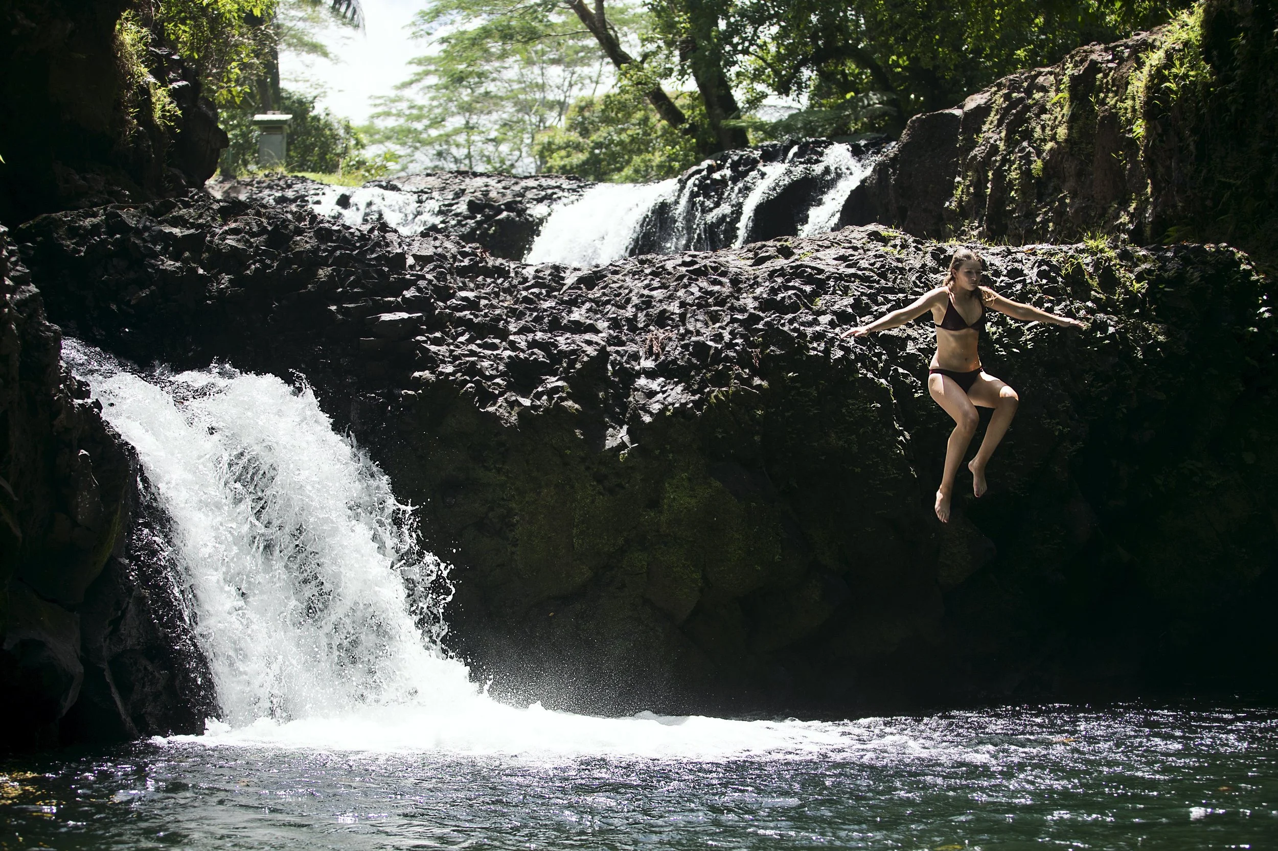A woman in a black bikini jumping off a large rock into a river near a waterfall surrounded by lush green trees.