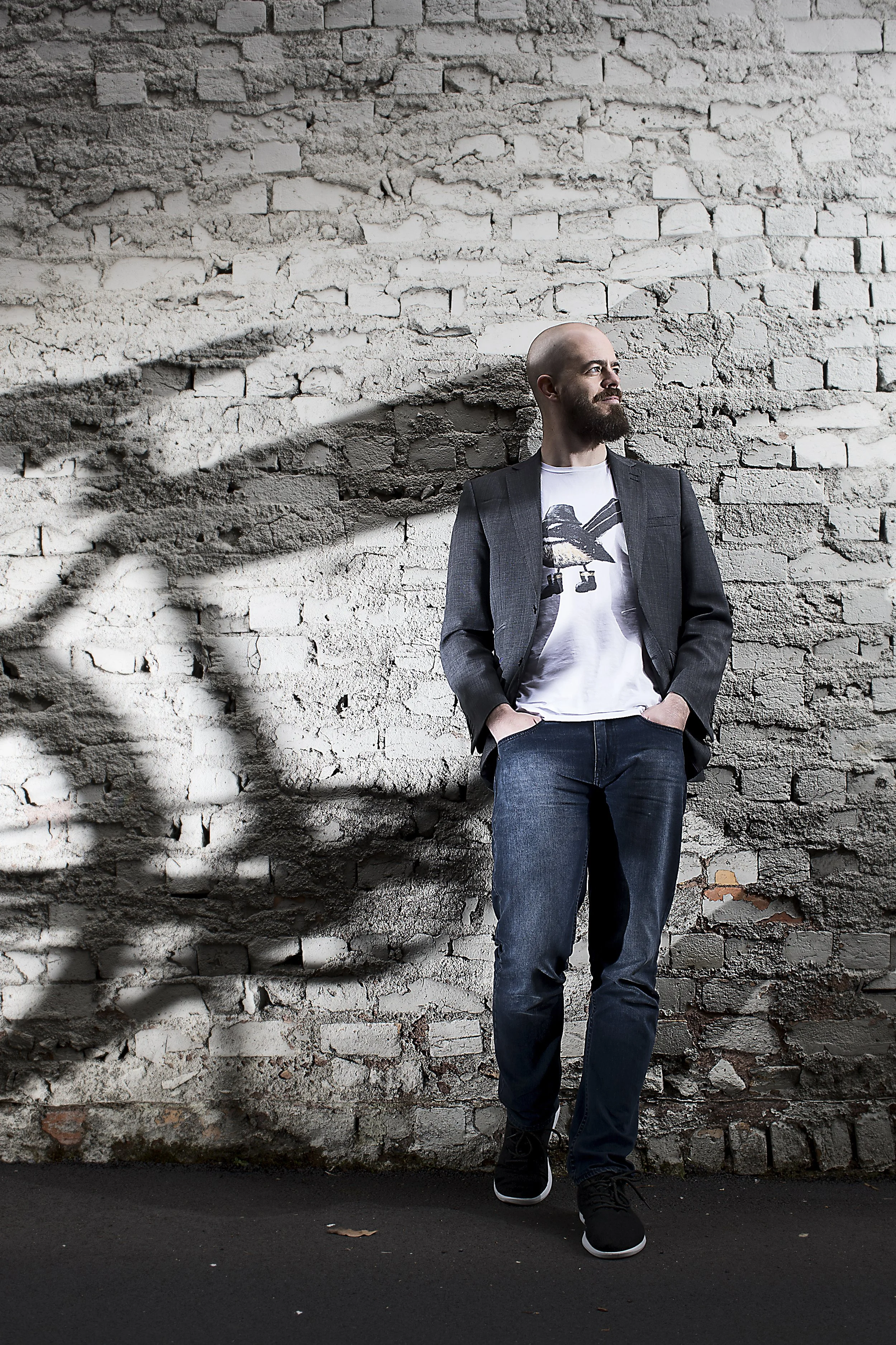 A man with a shaved head and beard is standing against a white brick wall with a shadow of tree branches cast on it. He is wearing a dark blazer, a white t-shirt with a graphic print, blue jeans, and black shoes. He has his hands in his pockets and i