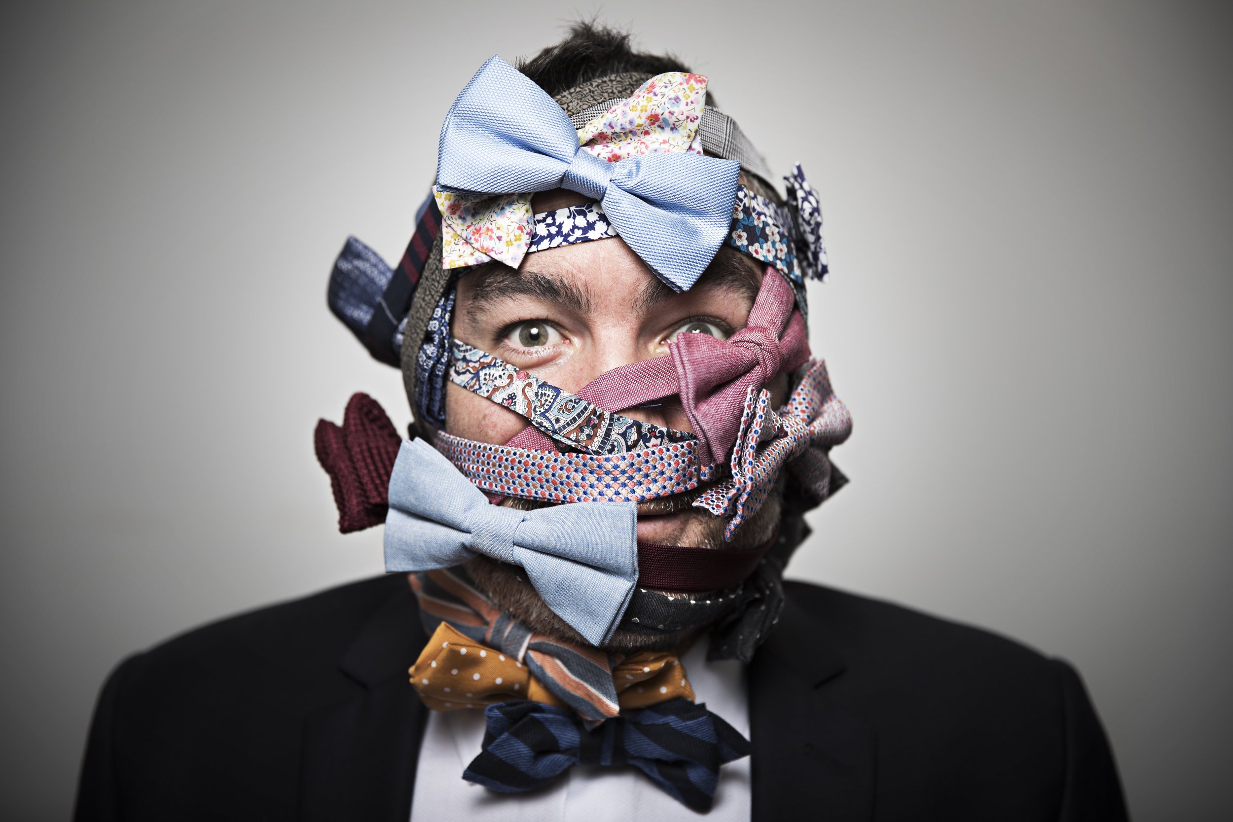 A man in a black suit with a white shirt is wearing multiple colorful ties in his face, tied around his head and face.