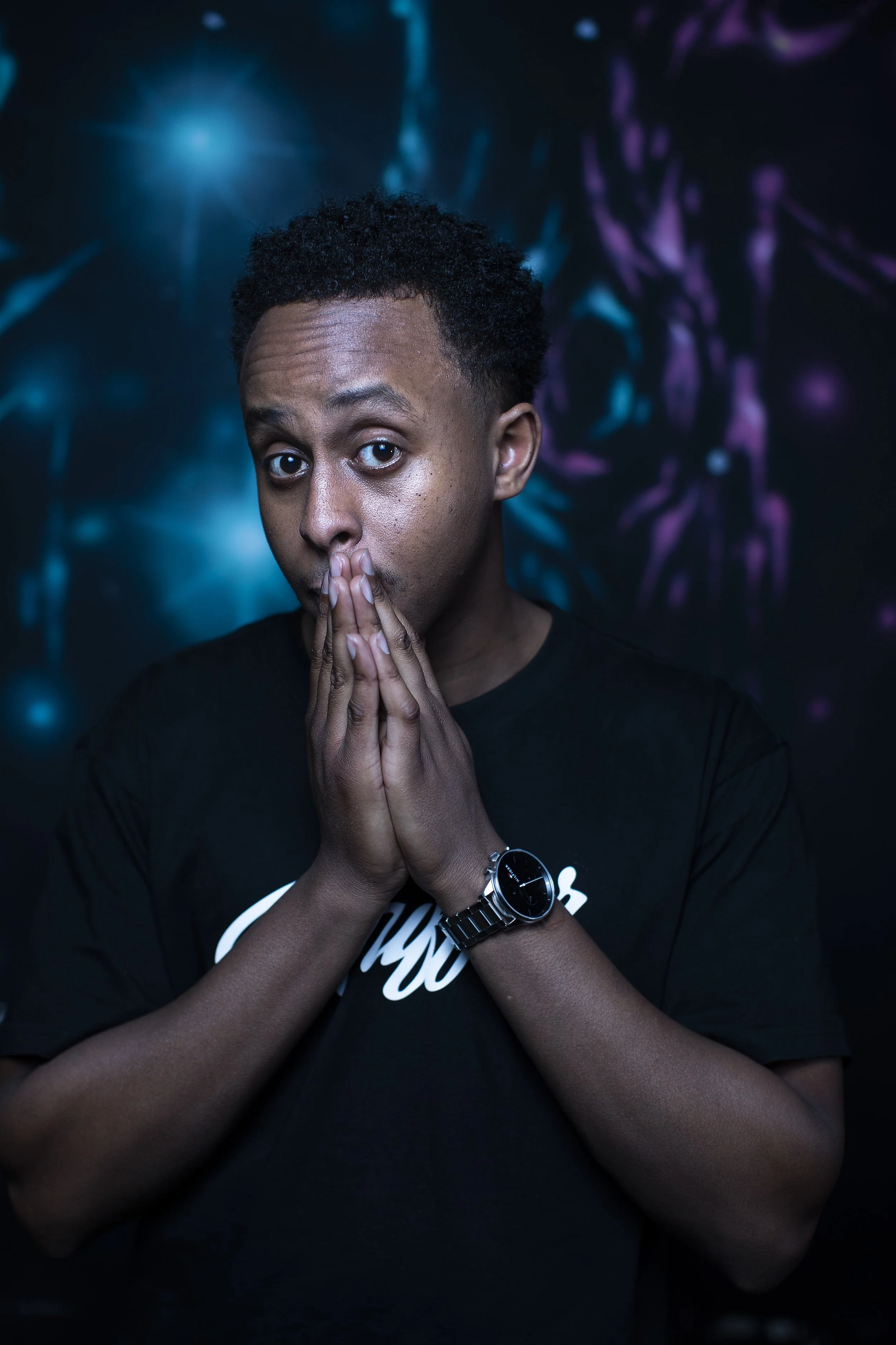 A young man with short curly hair, wearing a black t-shirt and a wristwatch, holds his hands near his mouth in a prayer-like gesture, looking directly at the camera with a surprised or contemplative expression. The background features a dark, abstrac