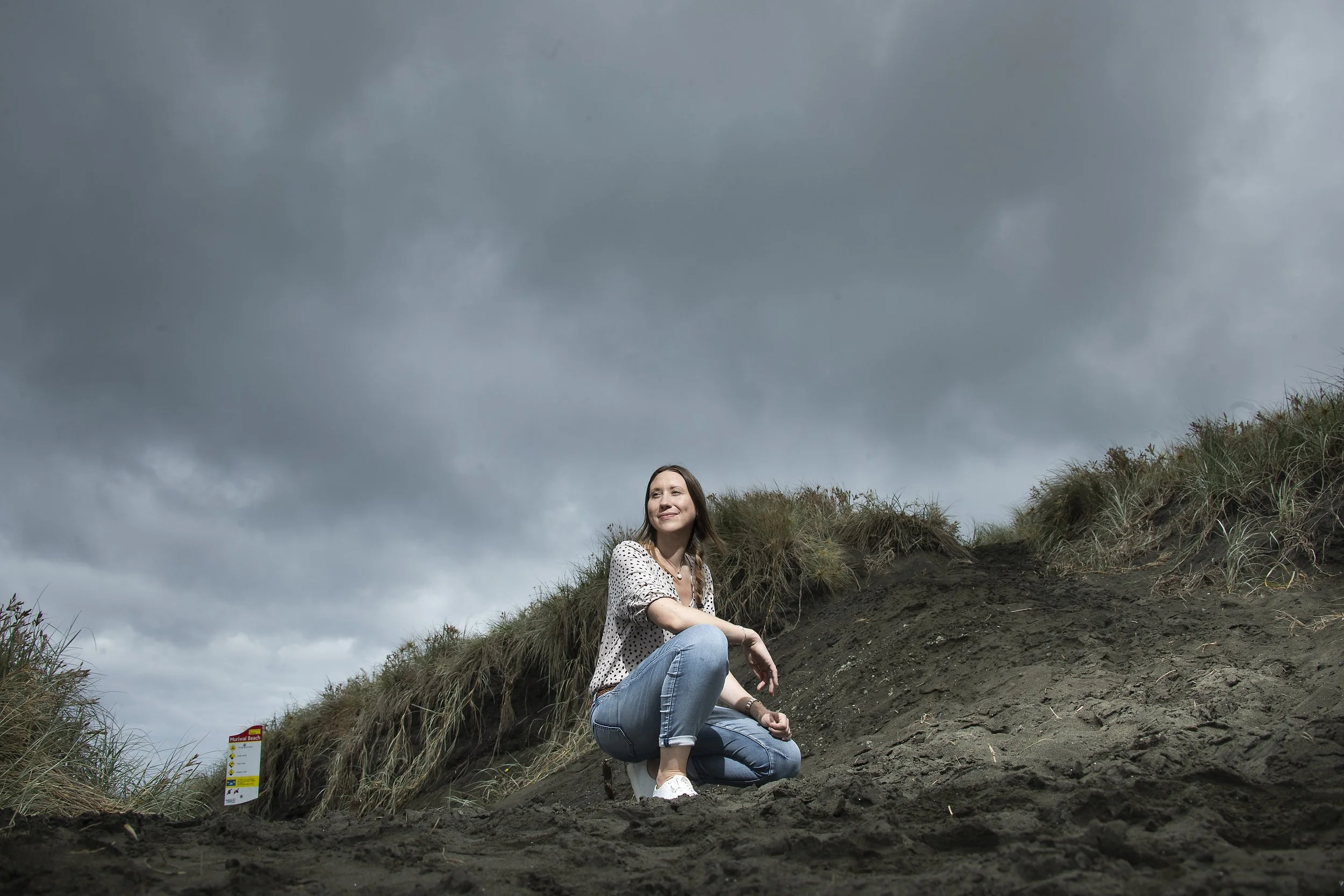 A woman crouching on dark sand at a beach with grassy dunes under a cloudy, overcast sky.