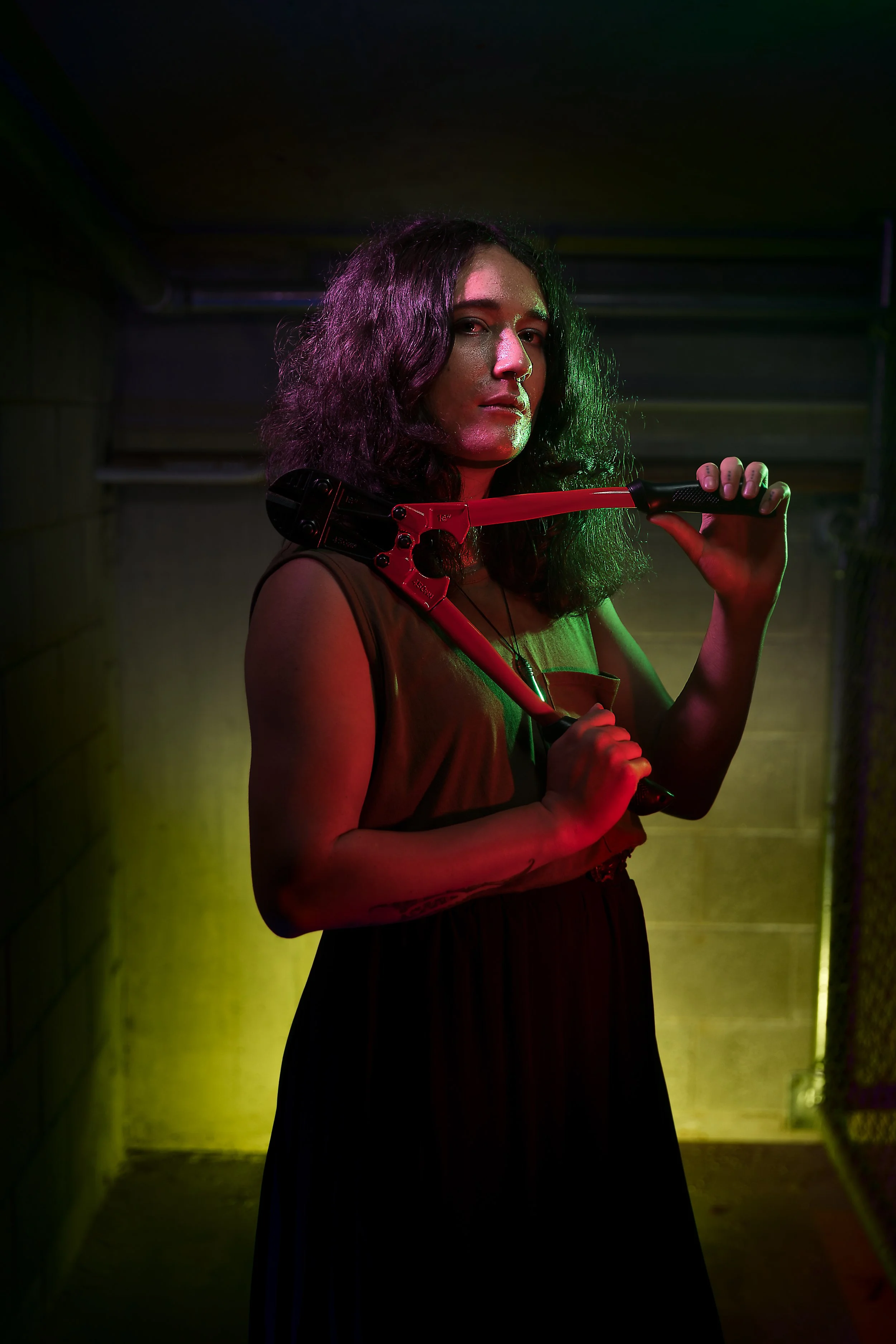 A woman with curly dark hair holds a red pipe wrench on her shoulder in a dimly lit room with green and yellow lighting.