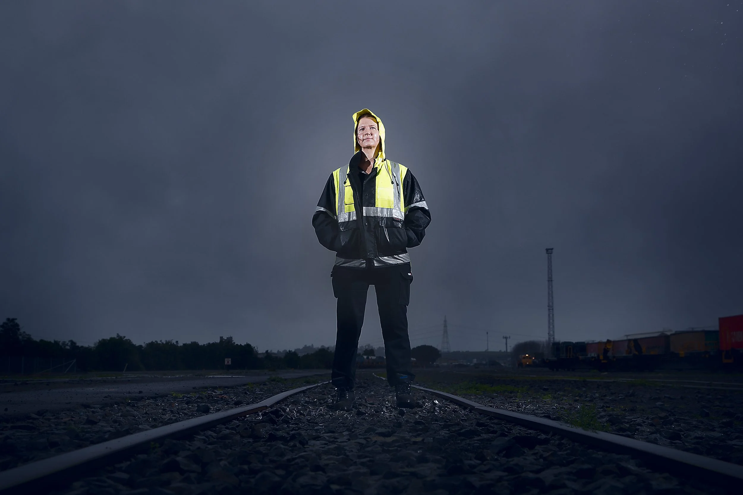 A person in a reflective jacket standing on railroad tracks at dusk or night, with a dark cloudy sky and distant train cars.