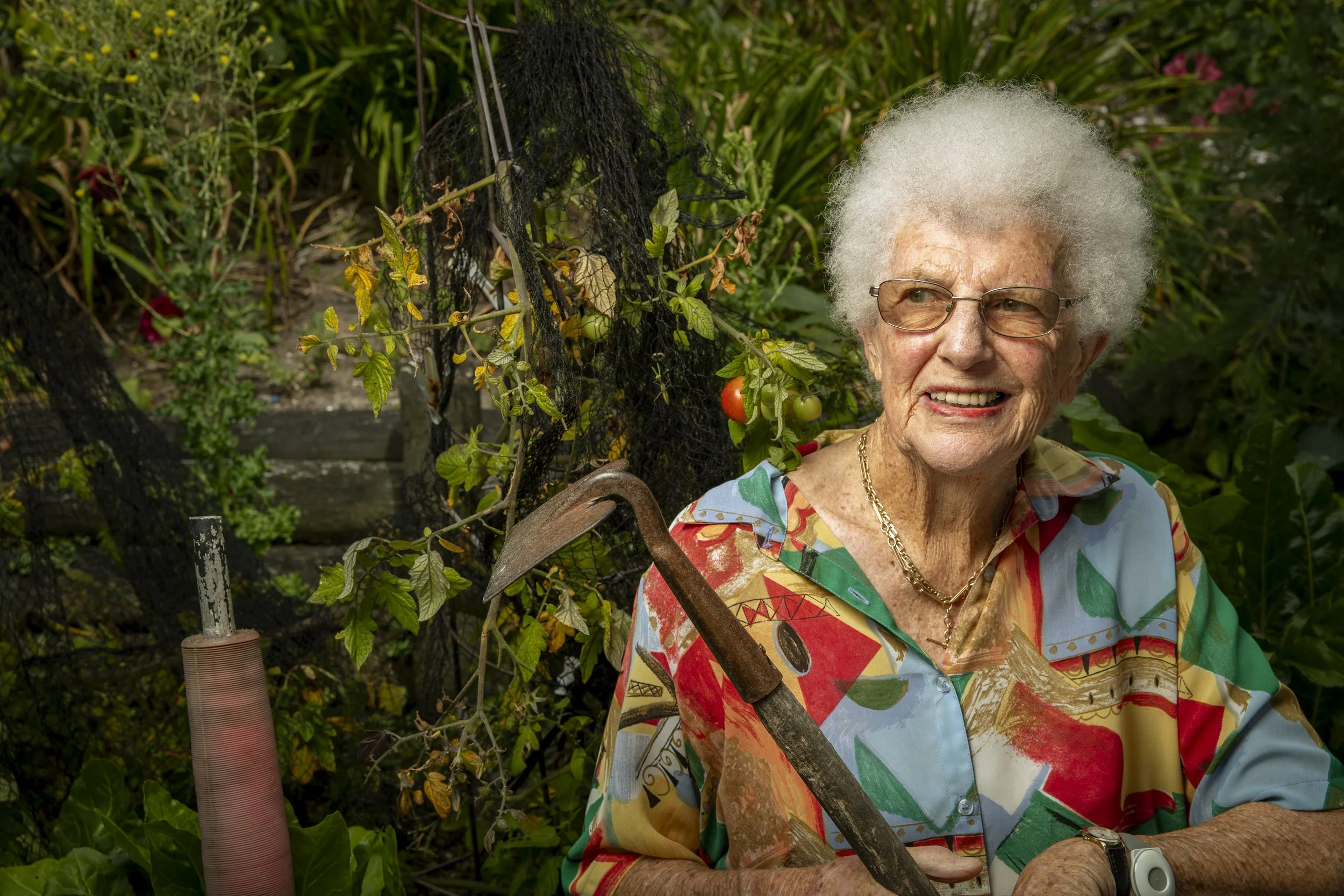 An elderly woman with white curly hair, wearing glasses, a colorful shirt, and a necklace, smiling while holding a gardening hoe in her garden surrounded by plants and tomatoes.