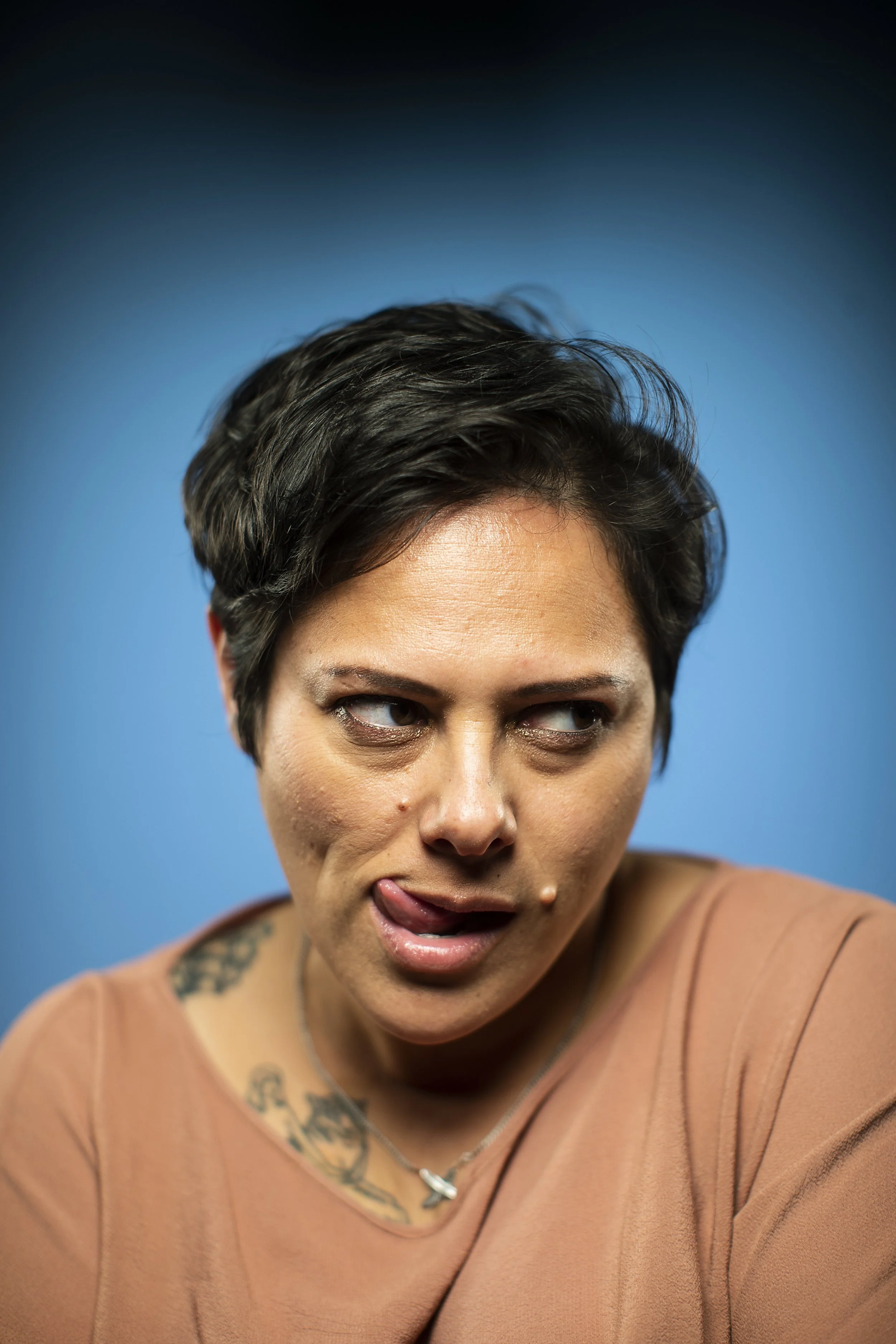 A woman with short dark hair making a playful face with her tongue out and eyes looking to the side, against a blue background.