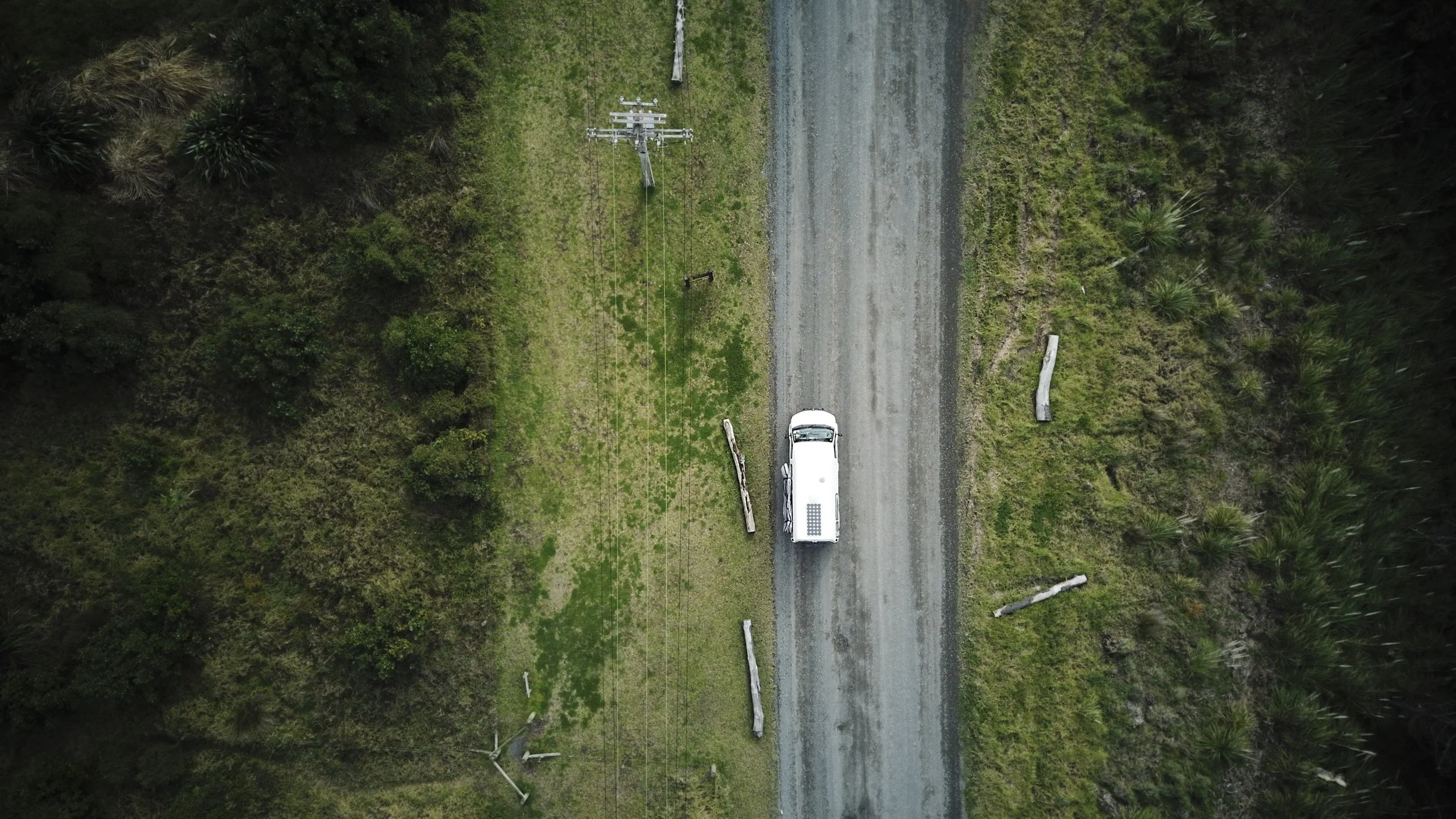 Aerial view of a white truck parked on a gravel road with damaged utility poles on either side, surrounded by green vegetation and trees.
