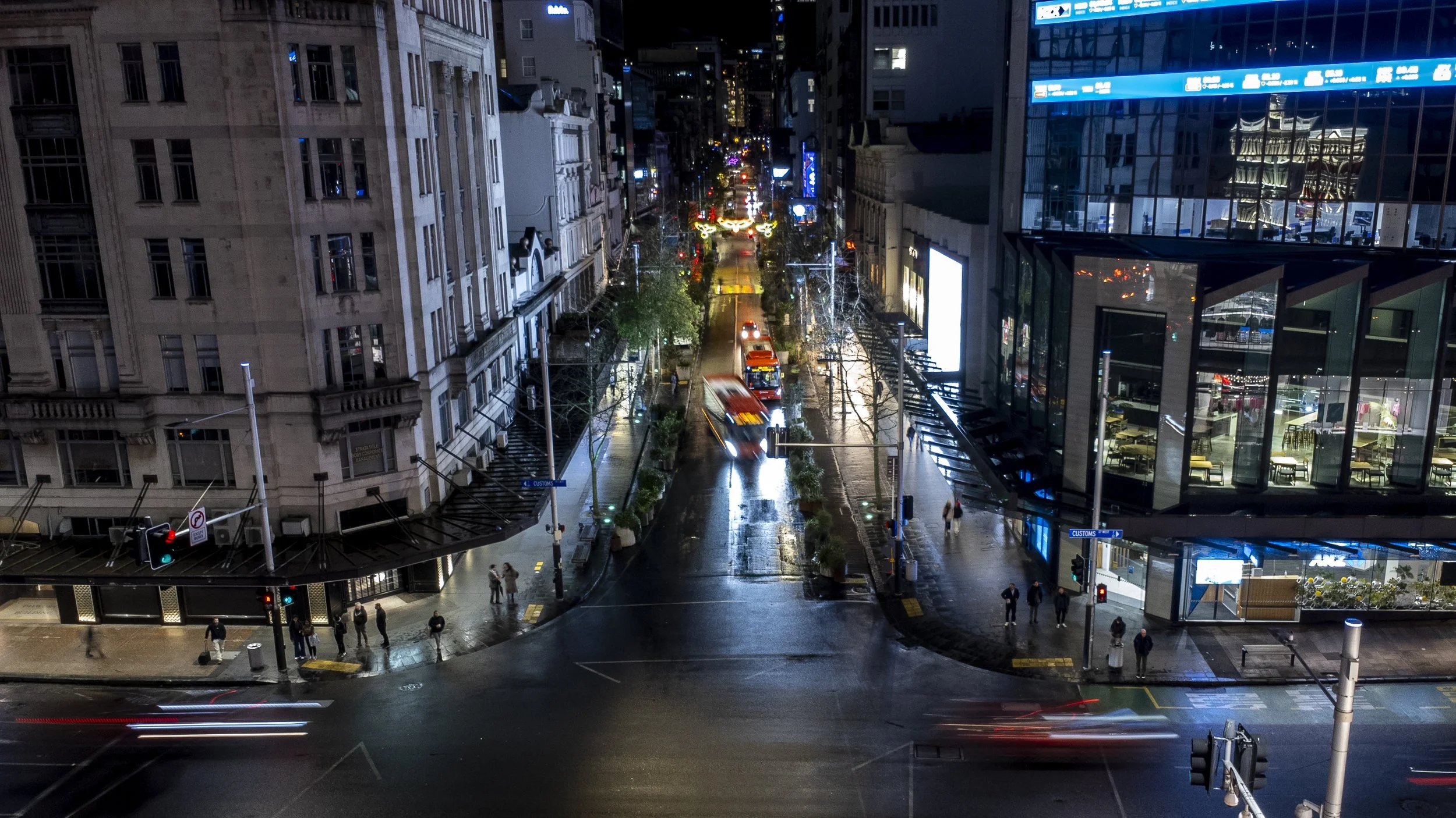Nighttime city street view with tall buildings, moving vehicles, pedestrians, and illuminated signs.