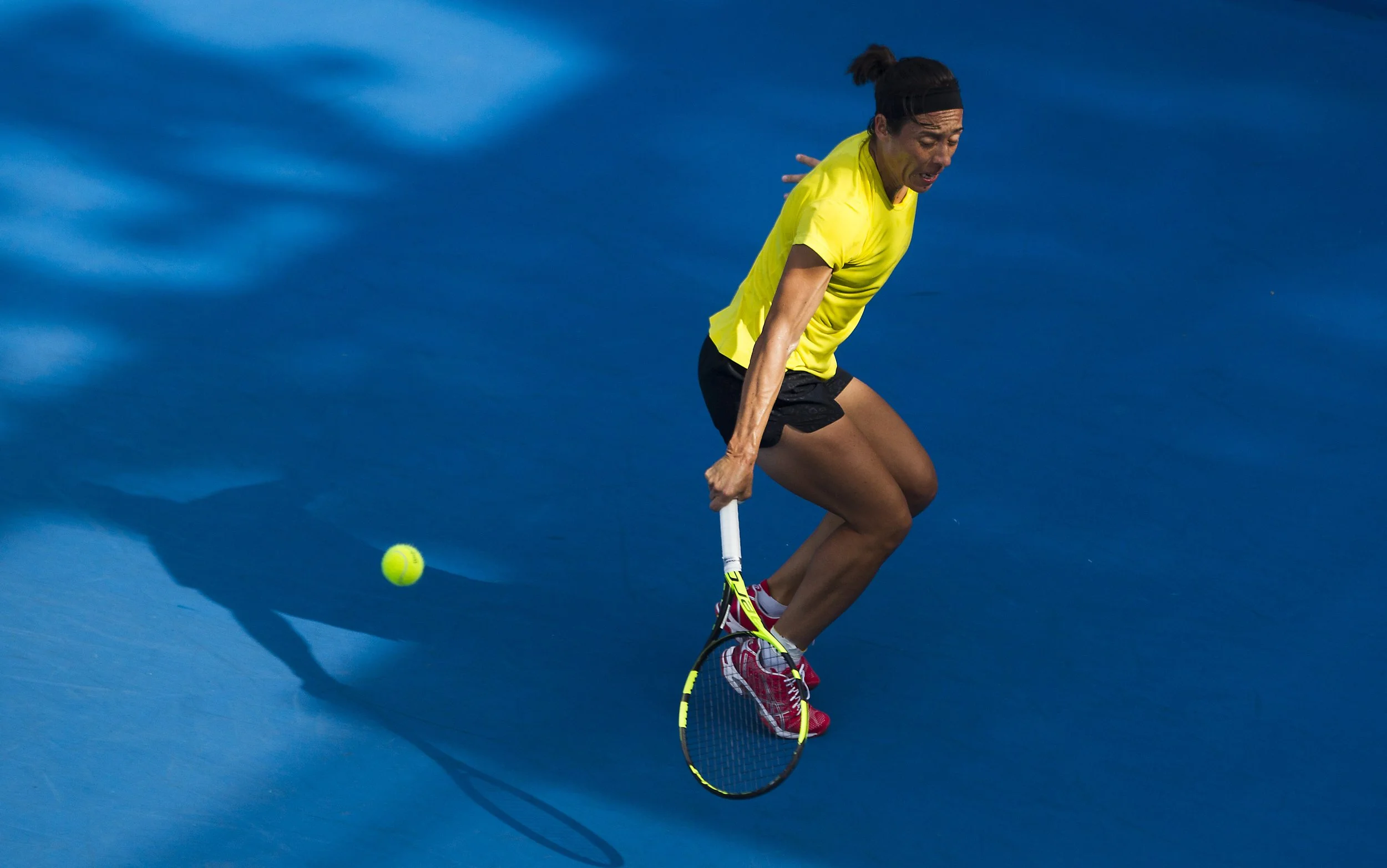 A male tennis player in a yellow shirt and black shorts on a blue court, preparing to hit a tennis ball with his racket.