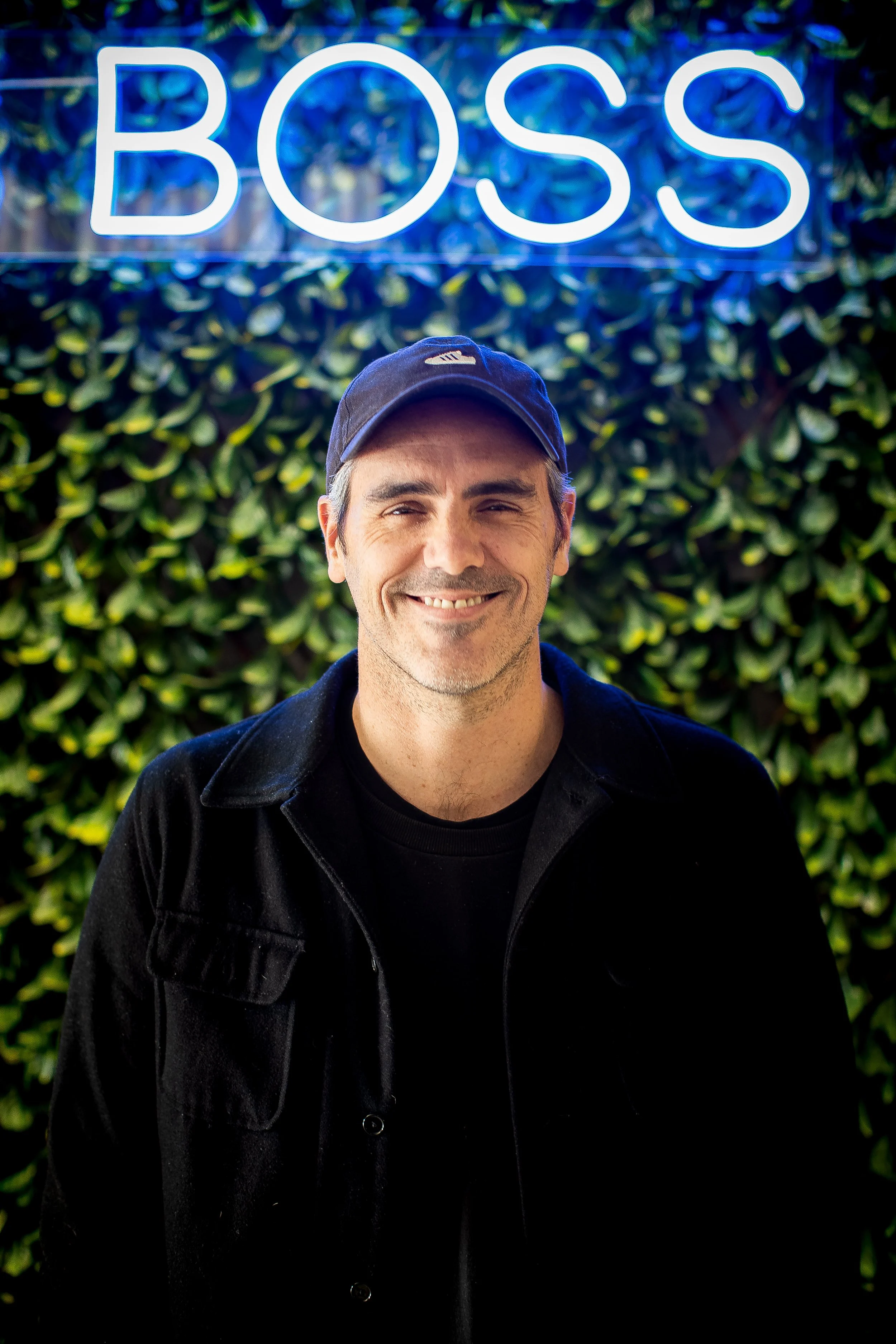 A man smiling in front of a foliage background with a neon sign above that says 'BOSS'.