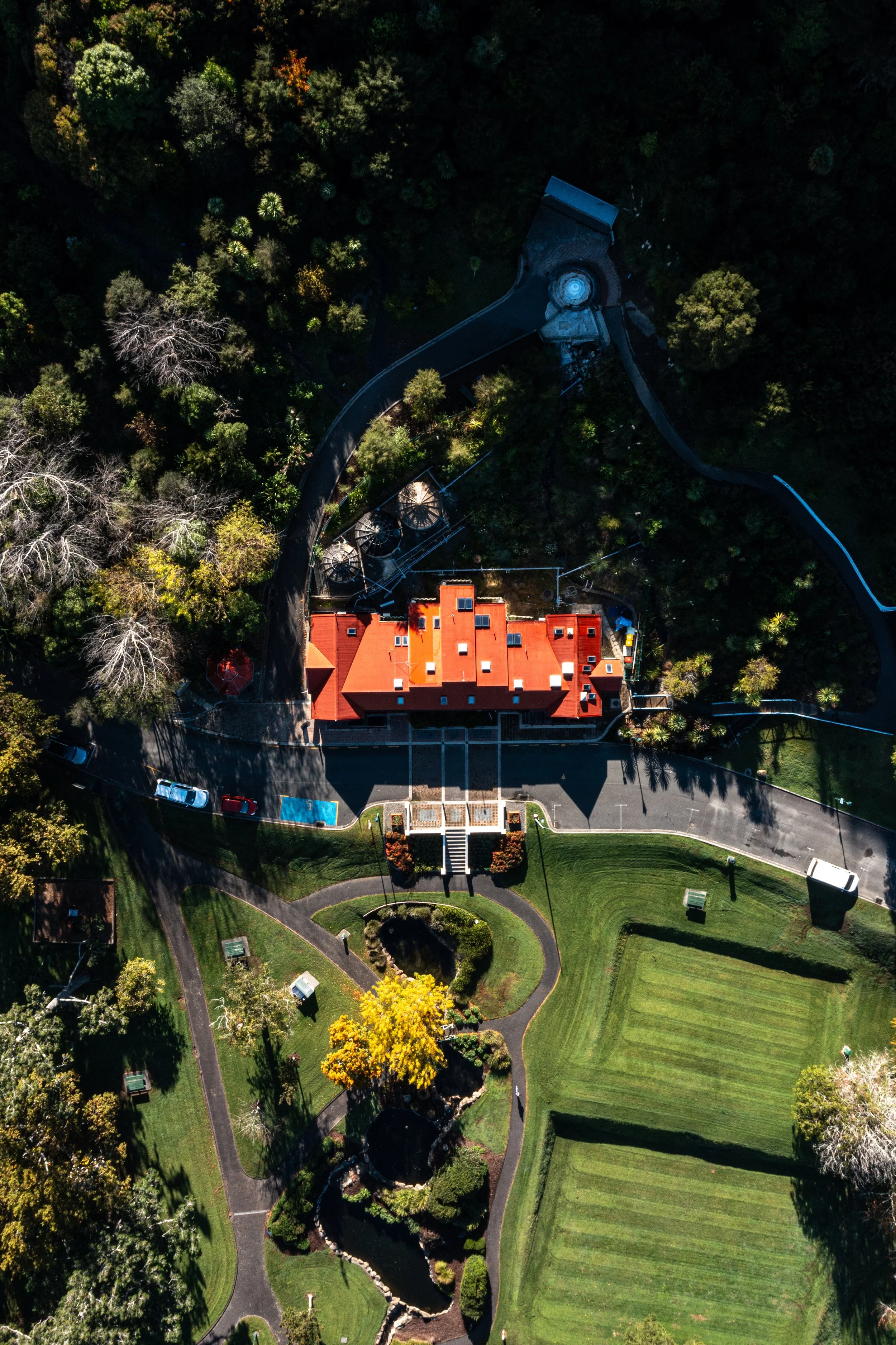 An aerial view of a building with a red roof, surrounded by a parking lot, pathways, trees, and landscaped gardens.