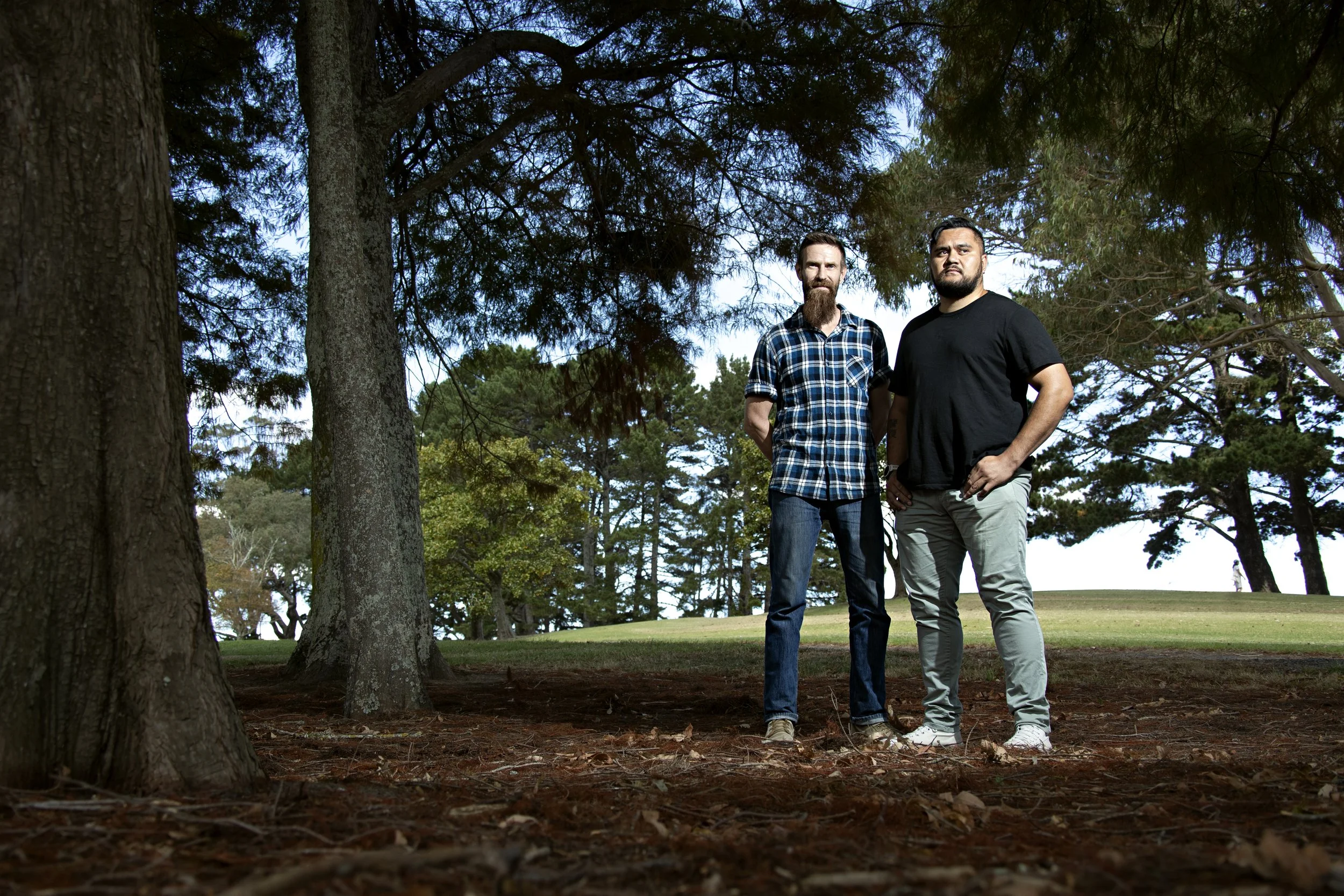 Two men standing outdoors in a park among tall trees, with one wearing a blue plaid shirt and the other a black t-shirt.