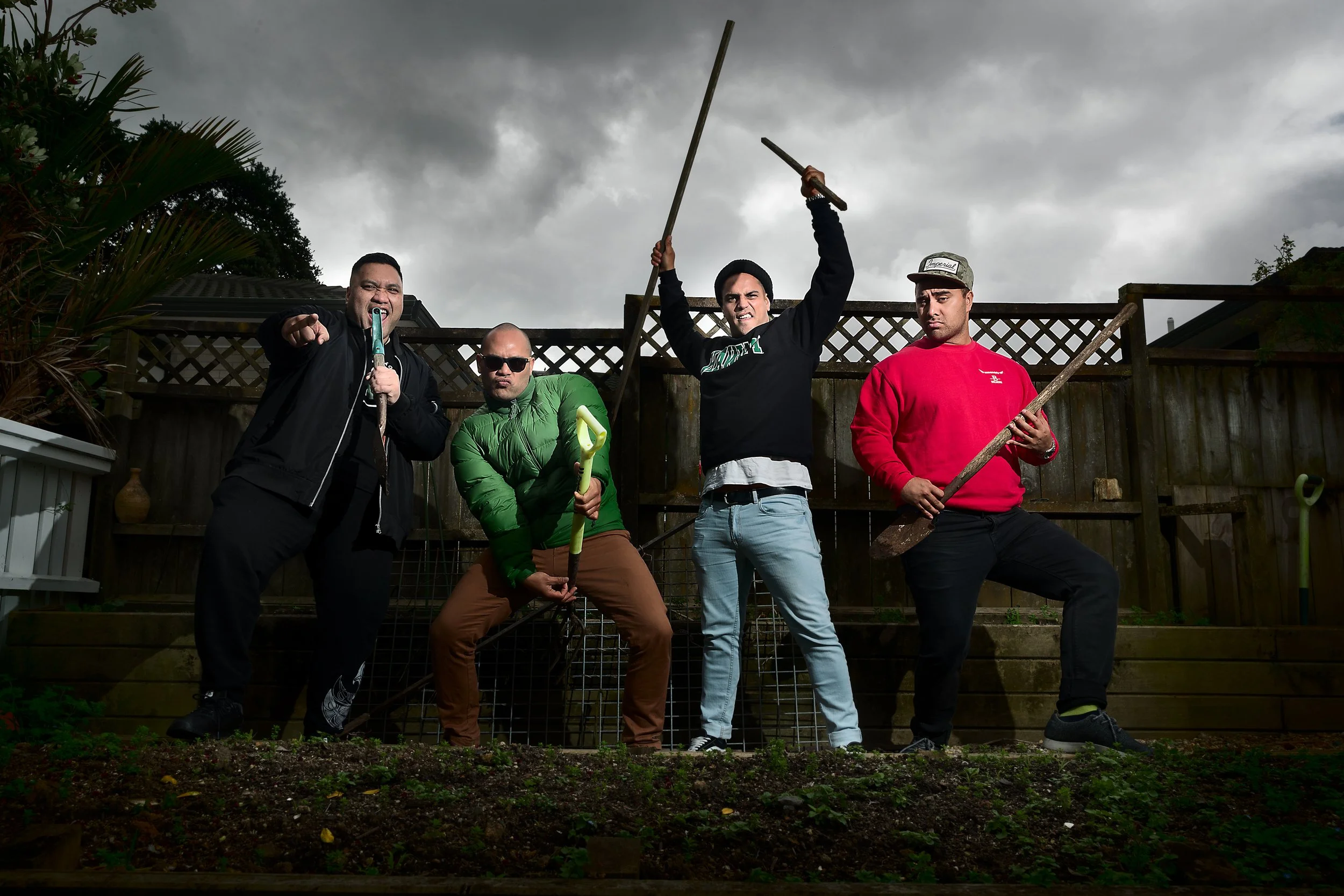 Four men with exaggerated poses holding gardening tools, standing in a backyard with a wooden fence and cloudy sky.