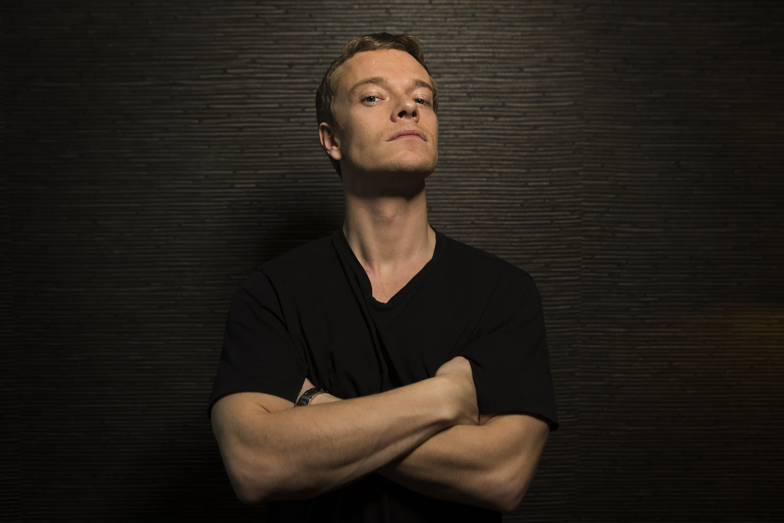 A young man with crossed arms looking confident, standing in front of a dark textured wall.