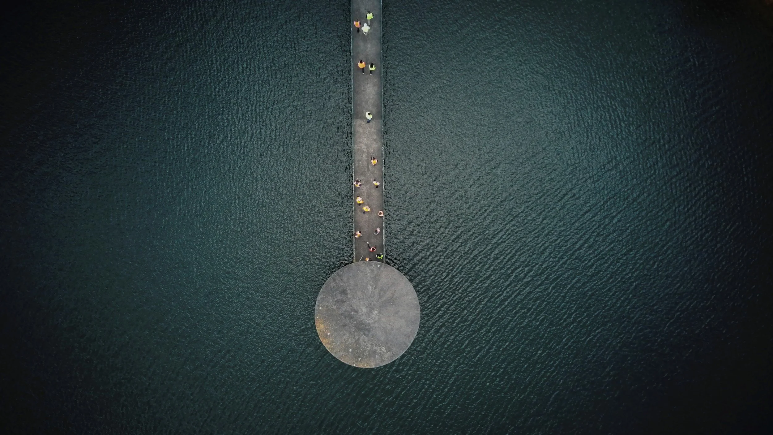 A circular concrete platform extends into the water from a pier, with people sitting and standing on the pier, viewed from above.