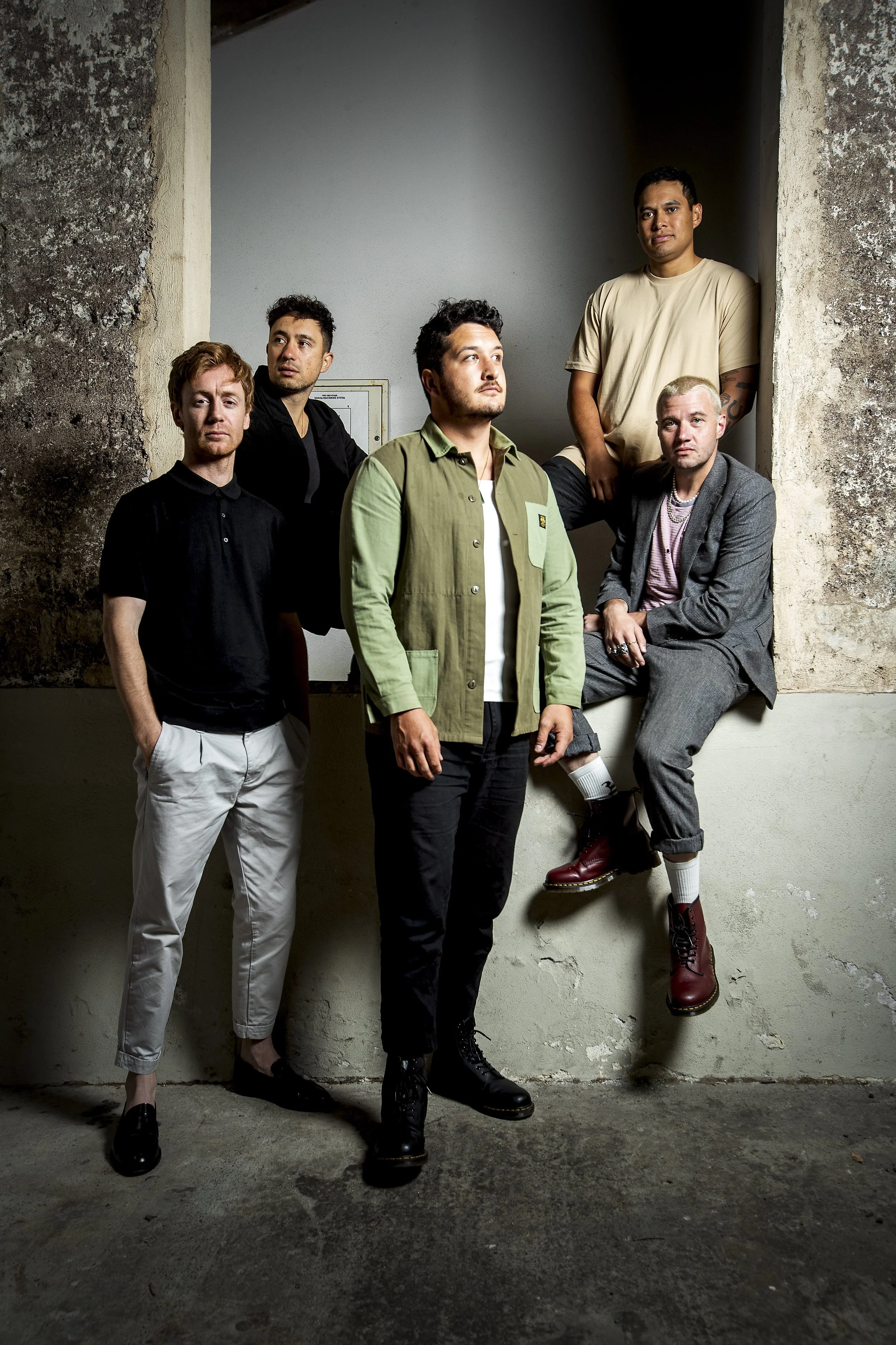 Group of five men standing and sitting in a gritty, industrial setting with weathered concrete walls. They are dressed in casual and stylish clothing, with some wearing jackets and boots.