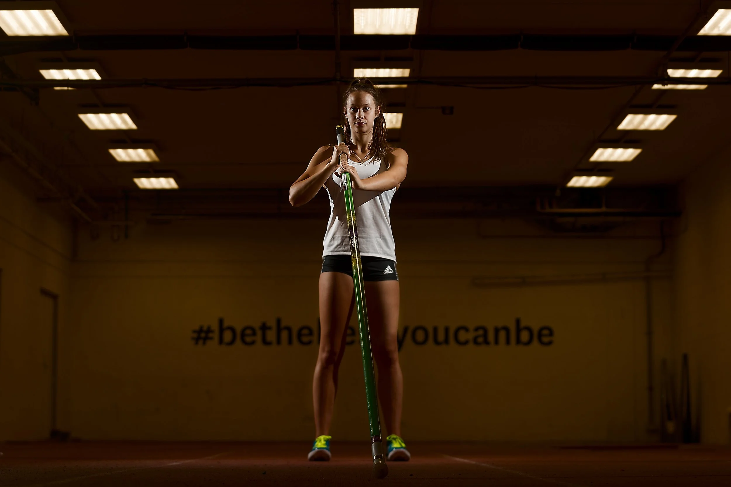A young woman standing in an indoor gymnasium, holding a pole vaulting pole, with a hashtag '#bethe' and words 'you can be' partially visible on the wall behind her.