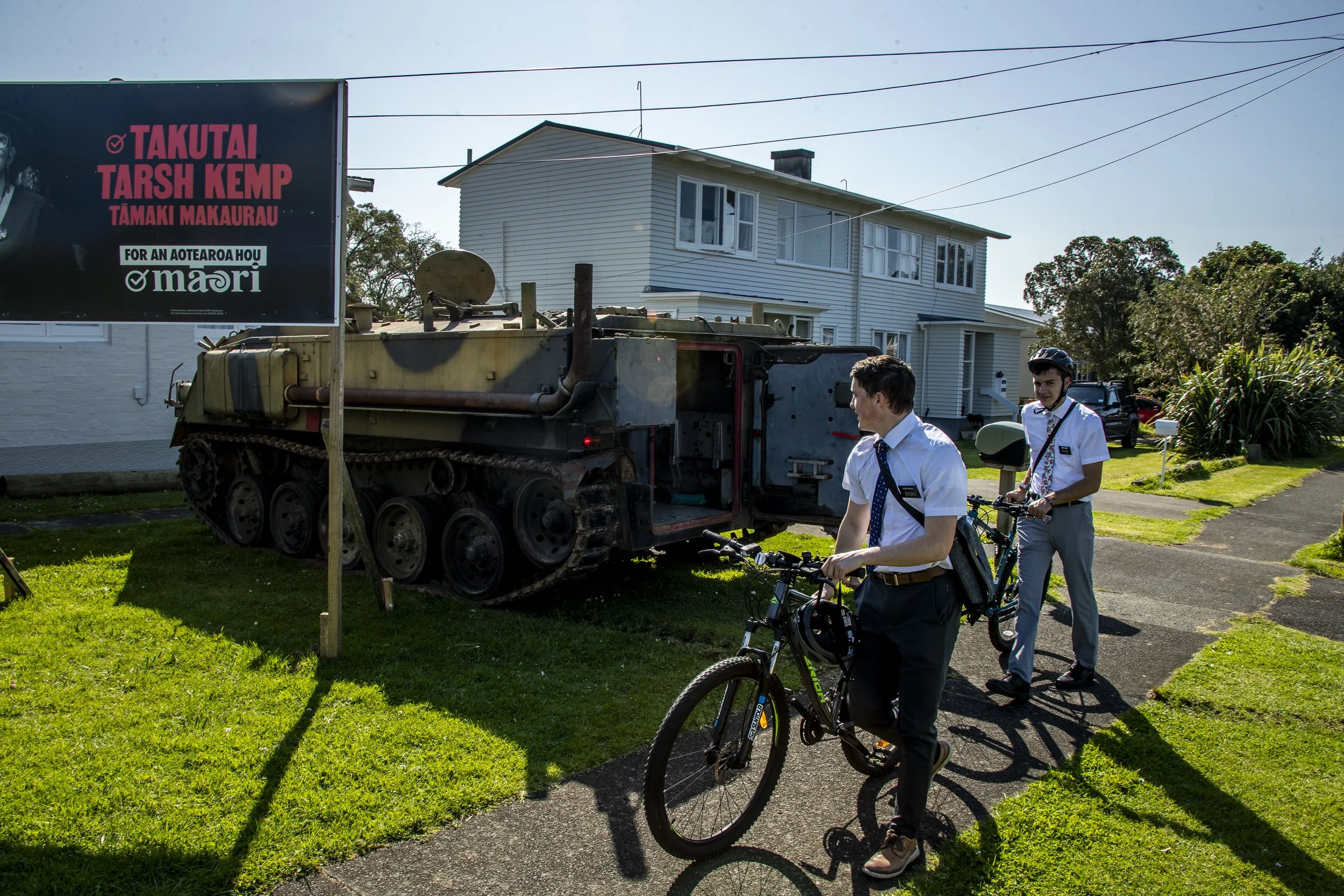 Two young men in school uniforms with bicycles observe a military tank displayed outdoors, next to a white house and a billboard with Māori text.
